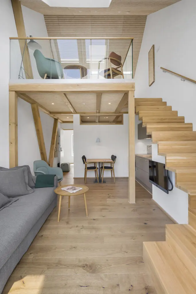 ferielejlighed danmark møllegården ringkøbing fjord En moderne, minimalistisk stue med trægulv og en mezzanin. Rummet byder på en grå sofa, et rundt sofabord i træ og en grøn lænestol. En spiseplads med bord og to stole er under mezzaninen, tilgængelig via trætrapper.