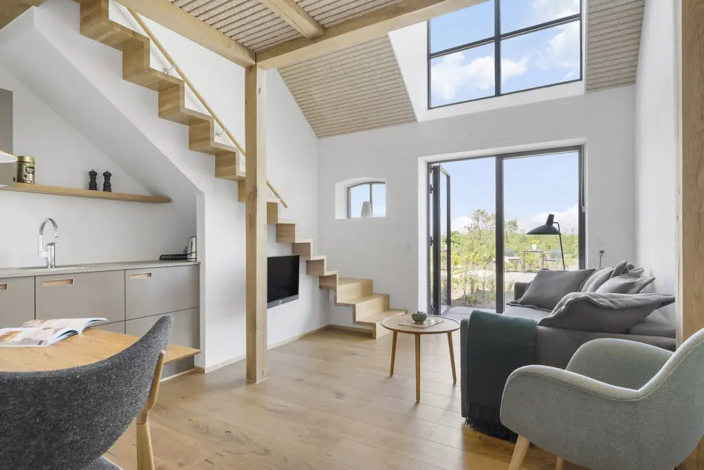 ferielejlighed danmark møllegården ringkøbing fjord Et moderne, minimalistisk opholdsrum med trægulve og højt til loftet. En trappe med åbne stiger fører til en hems. Værelset har en komfortabel sofa, en lænestol, et lille sofabord og store vinduer, der slipper naturligt lys ind. Køkkenafdeling til venstre.