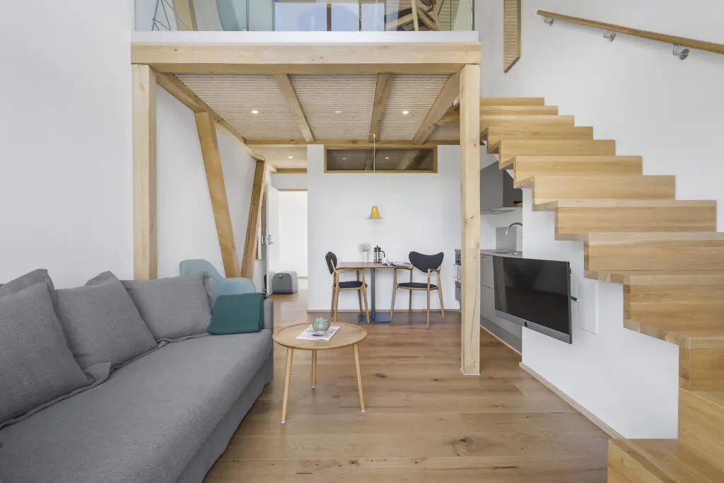 ferielejlighed danmark møllegården ringkøbing fjord Et moderne opholdsrum med trægulv og et åbent design. En grå sofa sidder til venstre, et sofabord af træ foran og en minimalistisk trætrappe uden stiger, der fører til et øverste niveau. Et lille spisebord med stole er bagerst.