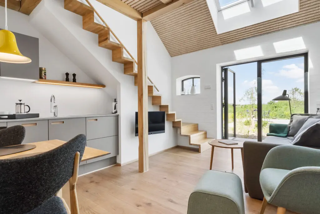 ferielejlighed danmark møllegården ringkøbing fjord Et moderne opholdsrum med en trappe med trætrapper, der fører ovenpå. Rummet har lyst trægulv, grå skabe, et lille køkkenområde og store glasdøre, der åbner ud til en have. Der er komfortable stole, en sofa og et rundt sofabord. Ovenlys oplyser rummet med naturligt lys.