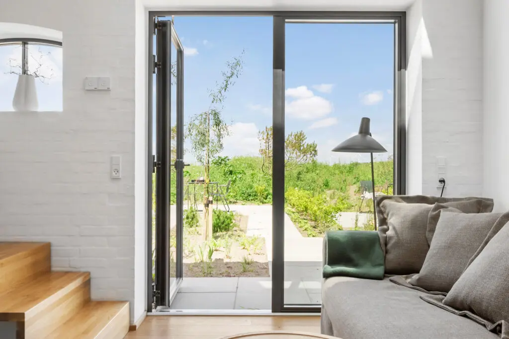 ferielejlighed danmark møllegården ringkøbing fjord Et lyst, moderne opholdsrum med en glasdør, der fører til en frodig have. Interiøret har en grå sofa med hynder, en elegant gulvlampe og trætrapper ved siden af en hvid murstensvæg. Naturligt lys oversvømmer rummet og skaber en luftig og indbydende atmosfære.