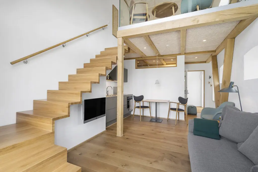 ferielejlighed danmark møllegården ringkøbing fjord En moderne hems med åbent koncept med trægulv og en trætrappe, der fører til et øverste niveau. Hovedområdet byder på en grå sofa, et væghængt tv, et lille køkken med grå skabe og et spisebord med to stole. Det øverste niveau har siddepladser og et glasrækværk.