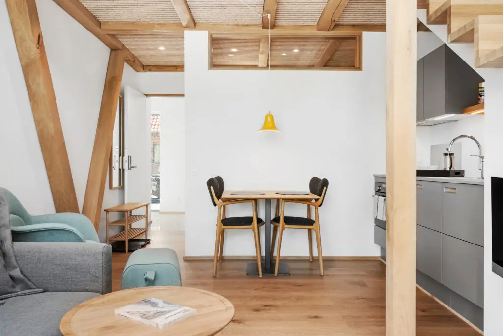 ferielejlighed danmark møllegården ringkøbing fjord Et hyggeligt moderne opholdsrum med et rundt sofabord i træ med en bog, en grå lænestol og en lille spiseplads med et bord, to stole og et hængende gult lys. Køkkenet har lækre grå skabe, og der er træbjælker i loftet.