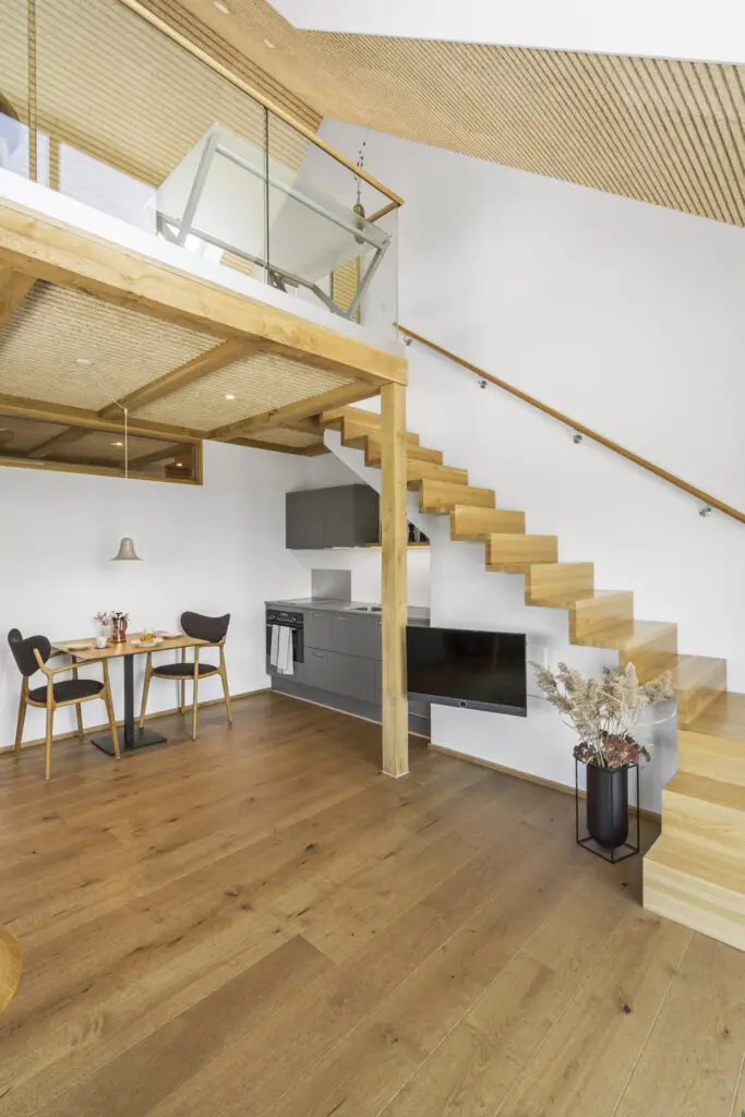ferielejlighed danmark møllegården ringkøbing fjord Et moderne, minimalistisk opholdsrum i åbent plan med trægulve, en flydende trætrappe med glasrækværk, et lille spisebord med to stole og et kompakt køkken med grå skabe. Ovenpå har en hems med træbjælker udsigt over området.