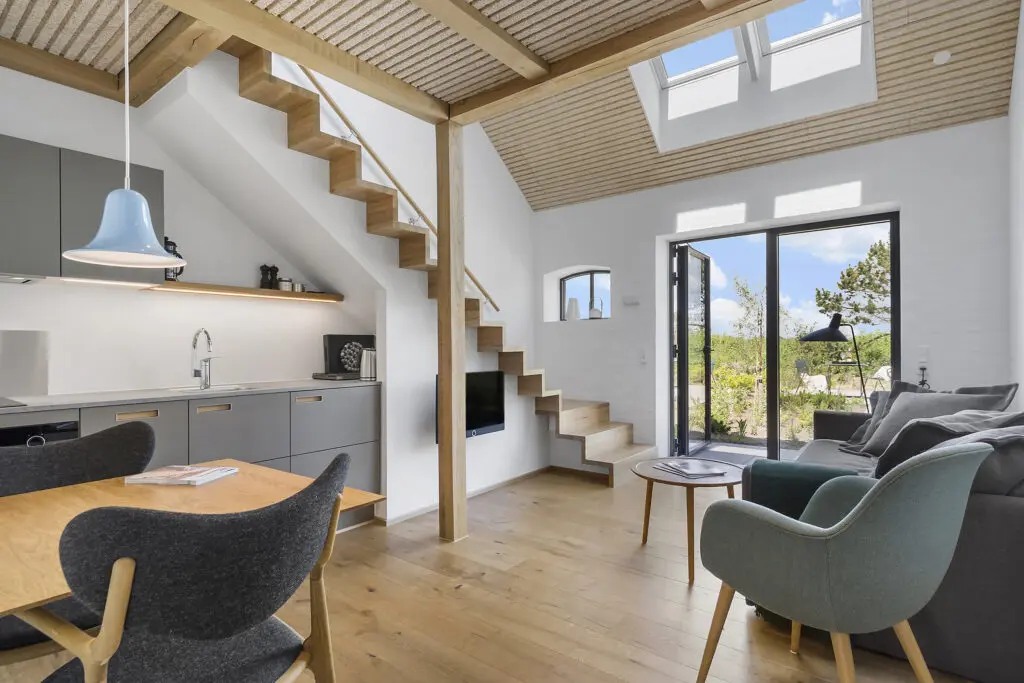 ferielejlighed danmark møllegården ringkøbing fjord Et moderne opholdsrum med åbent koncept med hems. Funktionerne omfatter trætrapper, ovenlys, store vinduer, en grå sofa, grønne og sorte loungestole, et spisebord i træ med sorte stole og et minimalistisk køkken med grå skabe. Lyse trægulve overalt.