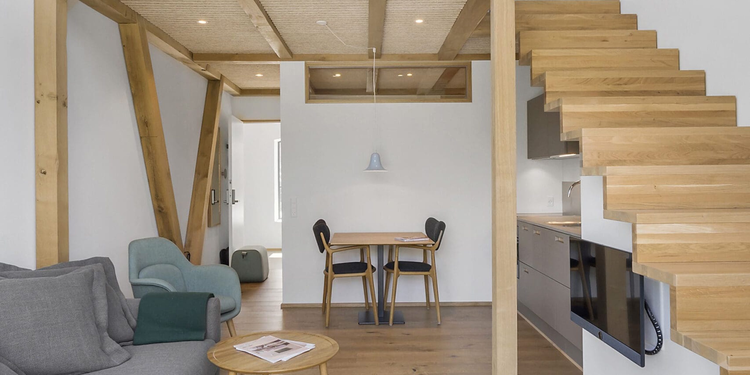 ferielejlighed danmark møllegården ringkøbing fjord En moderne, minimalistisk stue med trægulv og en mezzanin. Rummet byder på en grå sofa, et rundt sofabord i træ og en grøn lænestol. En spiseplads med bord og to stole er under mezzaninen, tilgængelig via trætrapper.