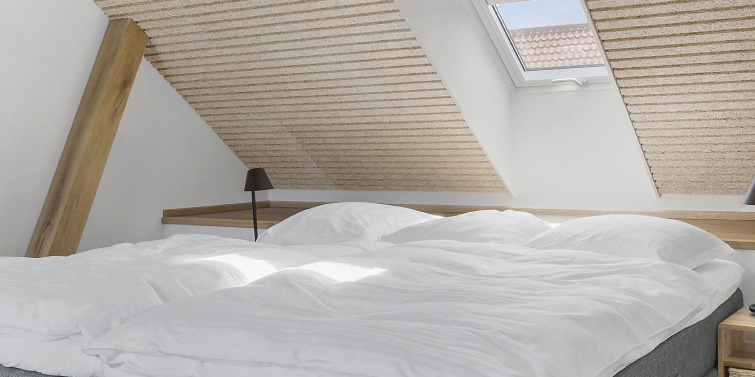 ferielejlighed danmark møllegården ringkøbing fjord Et hyggeligt loftsværelse med skrå loft og to ovenlysvinduer, med en pænt redet seng med grå polstret bund, hvidt sengetøj og et trænatbord med en lille sort lampe. Trægulvet komplementerer den lyse, moderne indretning.