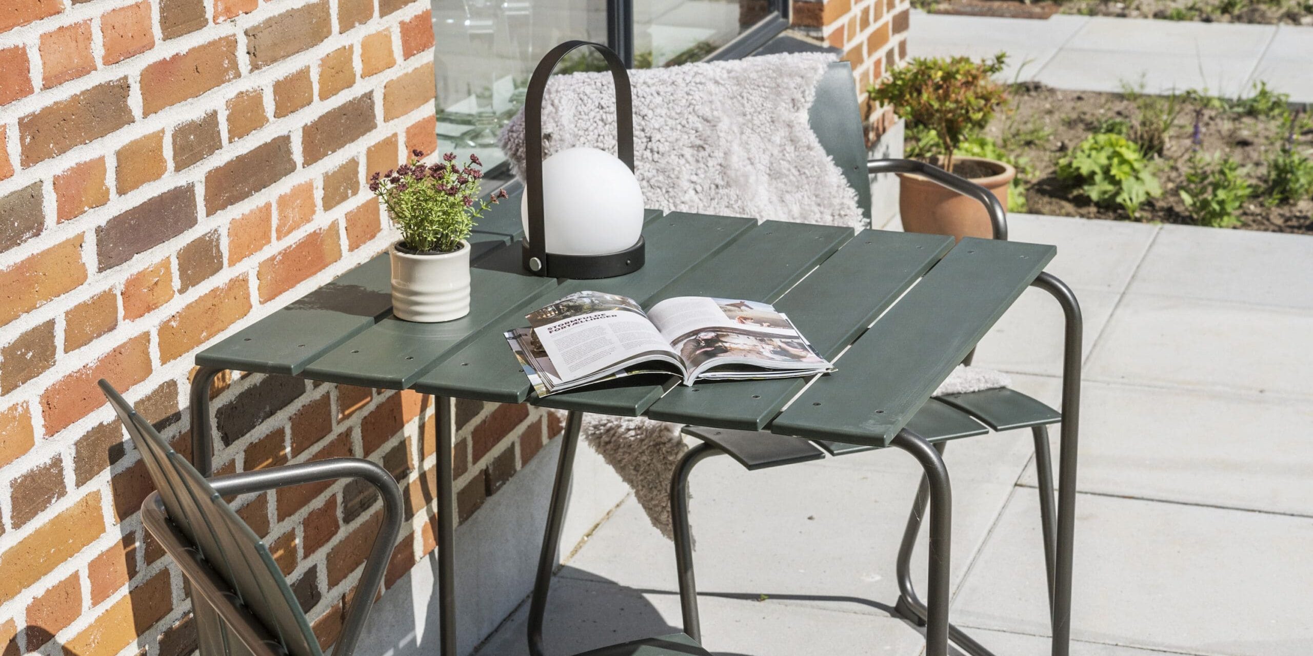 ferielejlighed danmark møllegården ringkøbing fjord En lille udendørs terrasse med et grønt bord og to stole mod en murstensvæg. Den ene stol har en hvid hynde draperet over ryggen. På bordet er der en åben bog, en lille potteplante og en lanterne med hvidt globuslys. Området er omgivet af planter og grønne områder.