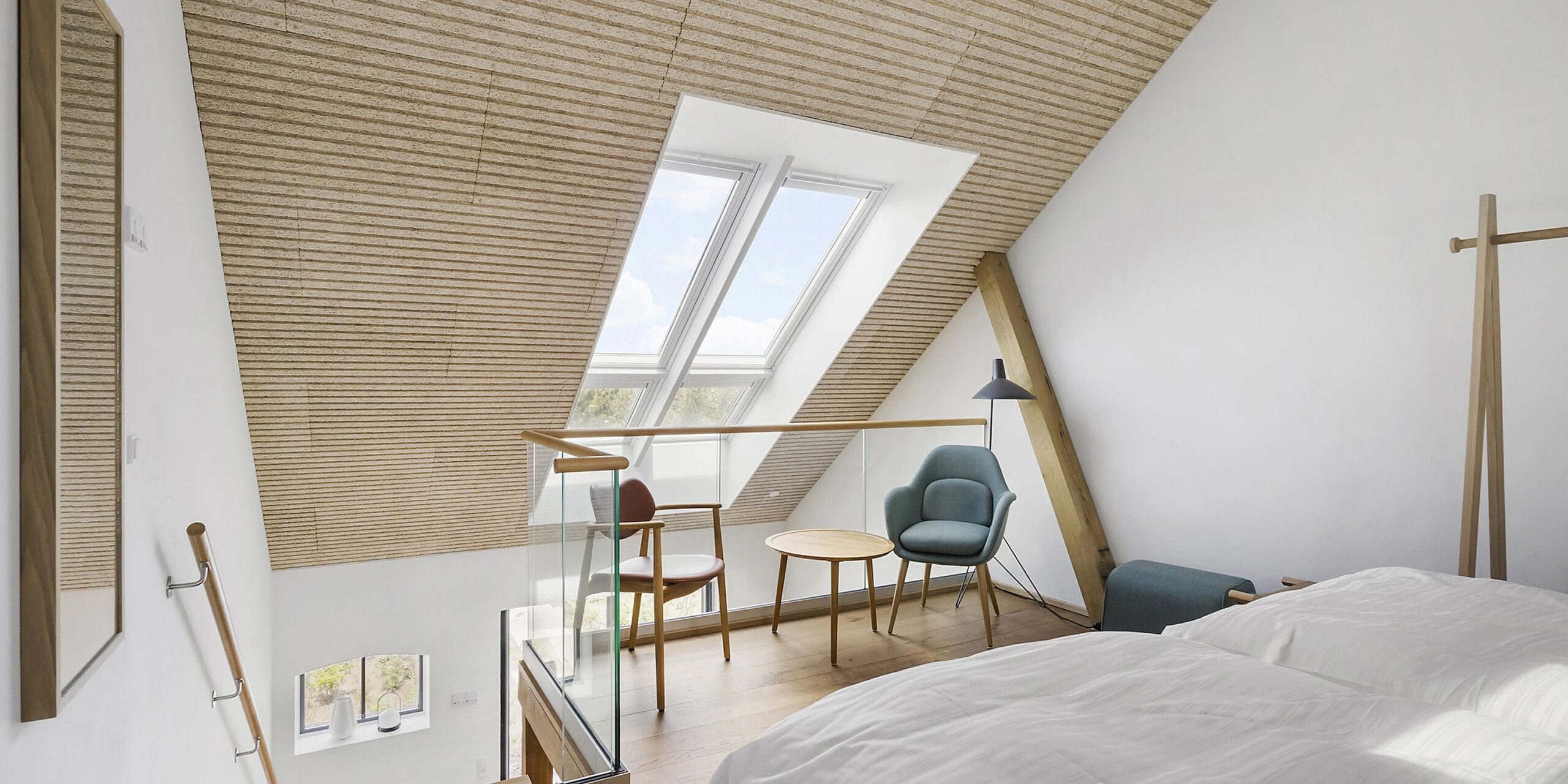 ferielejlighed danmark møllegården ringkøbing fjord Et moderne loftsværelse med skrå trælofter, ovenlysvinduer og et minimalistisk design. Værelset har en seng med hvidt sengetøj, to stole og et lille rundt bord ved vinduet, en gulvlampe i træ og et vægmonteret træspejl.