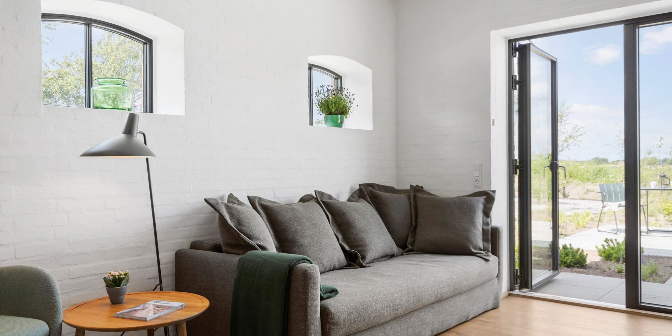 ferielejlighed danmark møllegården ringkøbing fjord En moderne stue med hvide murstensvægge og trægulve. Den har en grå sofa med flere hynder, et rundt sofabord i træ, en grøn lænestol, en gulvlampe og to små planter i vindueskarme. Glasdøren åbner til en naturskøn udendørs udsigt.