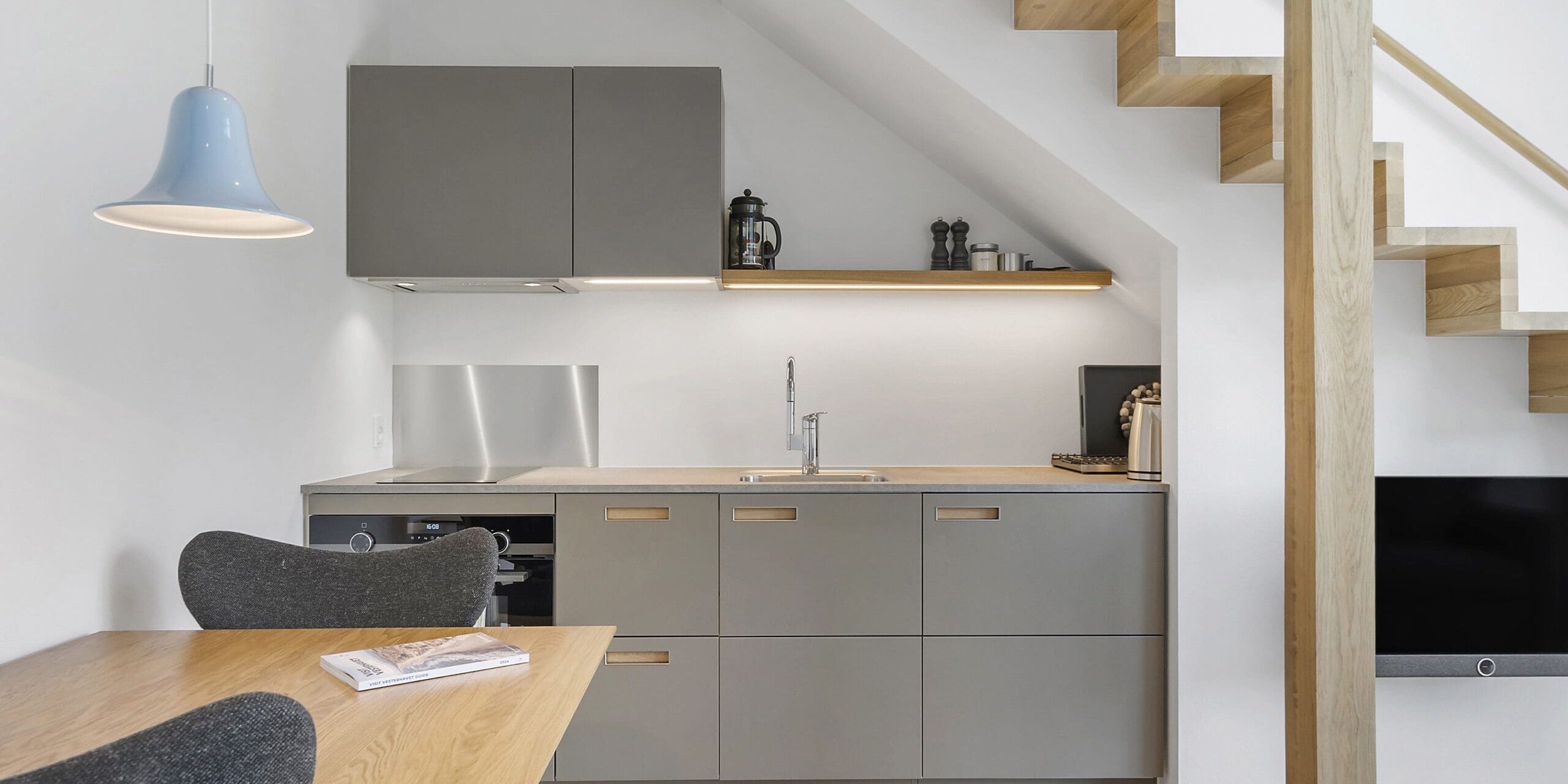 ferielejlighed danmark møllegården ringkøbing fjord Et moderne, minimalistisk køkken under en trætrappe. Køkkenet byder på slanke, grå skabe med integrerede hvidevarer, bordplade med vask og hylder. Tilstødende er en lille spiseplads med et træbord, sorte stole og en pendel ovenover.