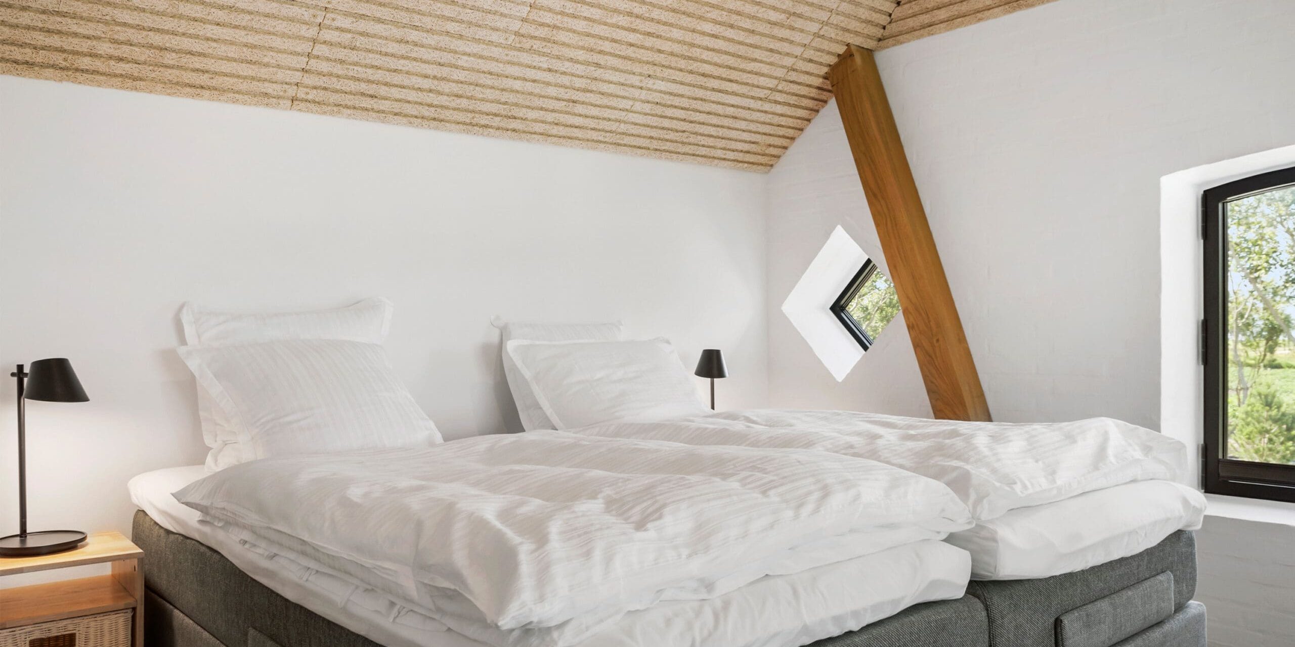 ferielejlighed danmark møllegården ringkøbing fjord Et hyggeligt soveværelse med et skrå loft med træpaneler og hvide vægge har to enkeltsenge med sprødt hvidt sengetøj placeret sammen til en dobbeltseng. To små vinduer, et kvadratisk og et rektangulært, giver naturligt lys ind. Et natbord i træ med en lampe er ved siden af en seng.
