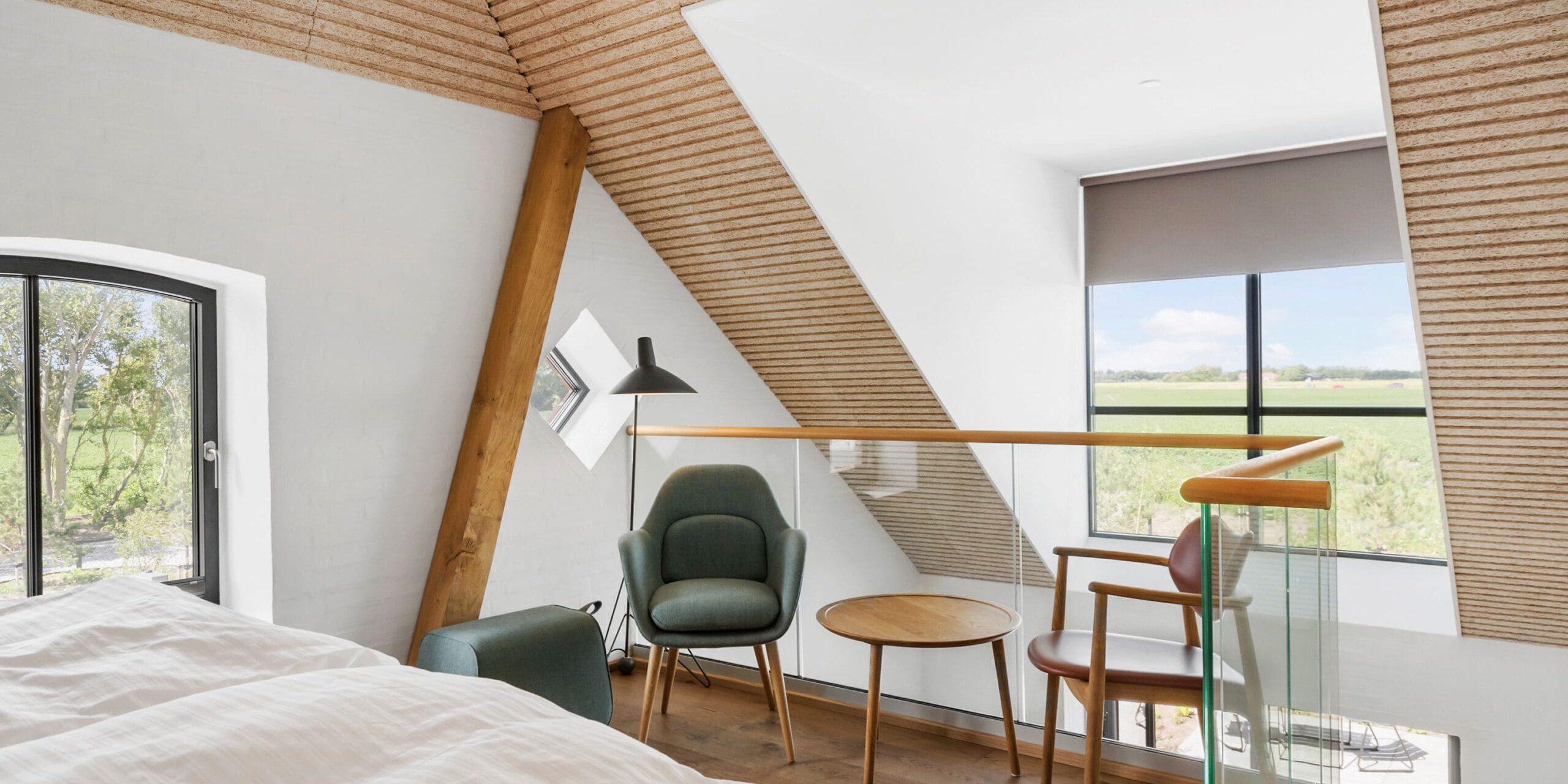 ferielejlighed danmark møllegården ringkøbing fjord En hyggelig hems med skrå træloft, med en seng med hvidt sengetøj, to moderne lænestole, et lille rundt træbord og et stort vindue med en naturskøn udsigt over grønne marker udenfor. Lyset filtrerer ind gennem vinduerne og oplyser rummet.