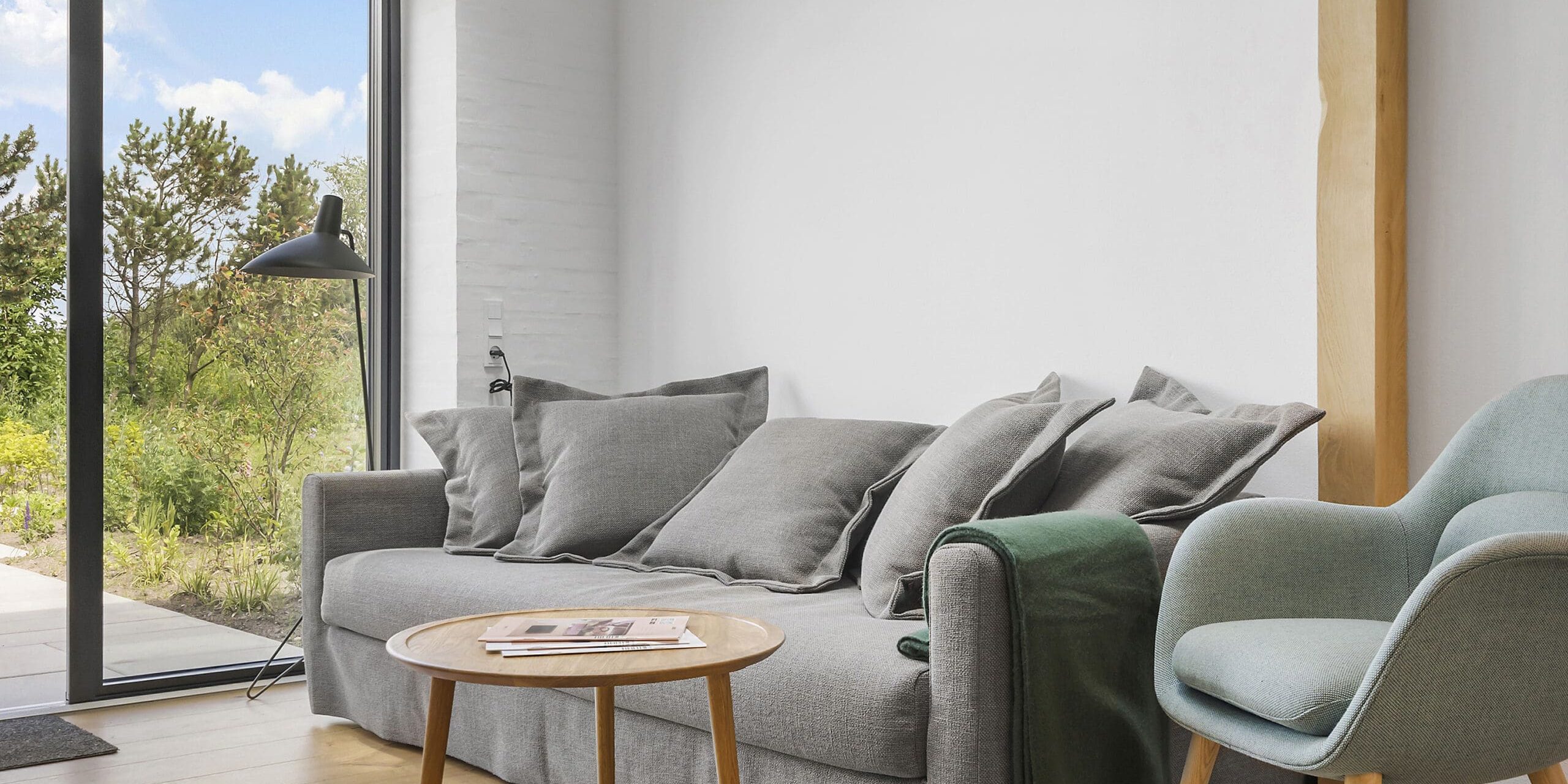 ferielejlighed danmark møllegården ringkøbing fjord En moderne stue med en grå sofa prydet med puder og et grønt tæppe, et rundt sofabord i træ og en blå lænestol. En sort gulvlampe står ved et stort vindue, der giver udsigt til grønne områder og en klar blå himmel udenfor.