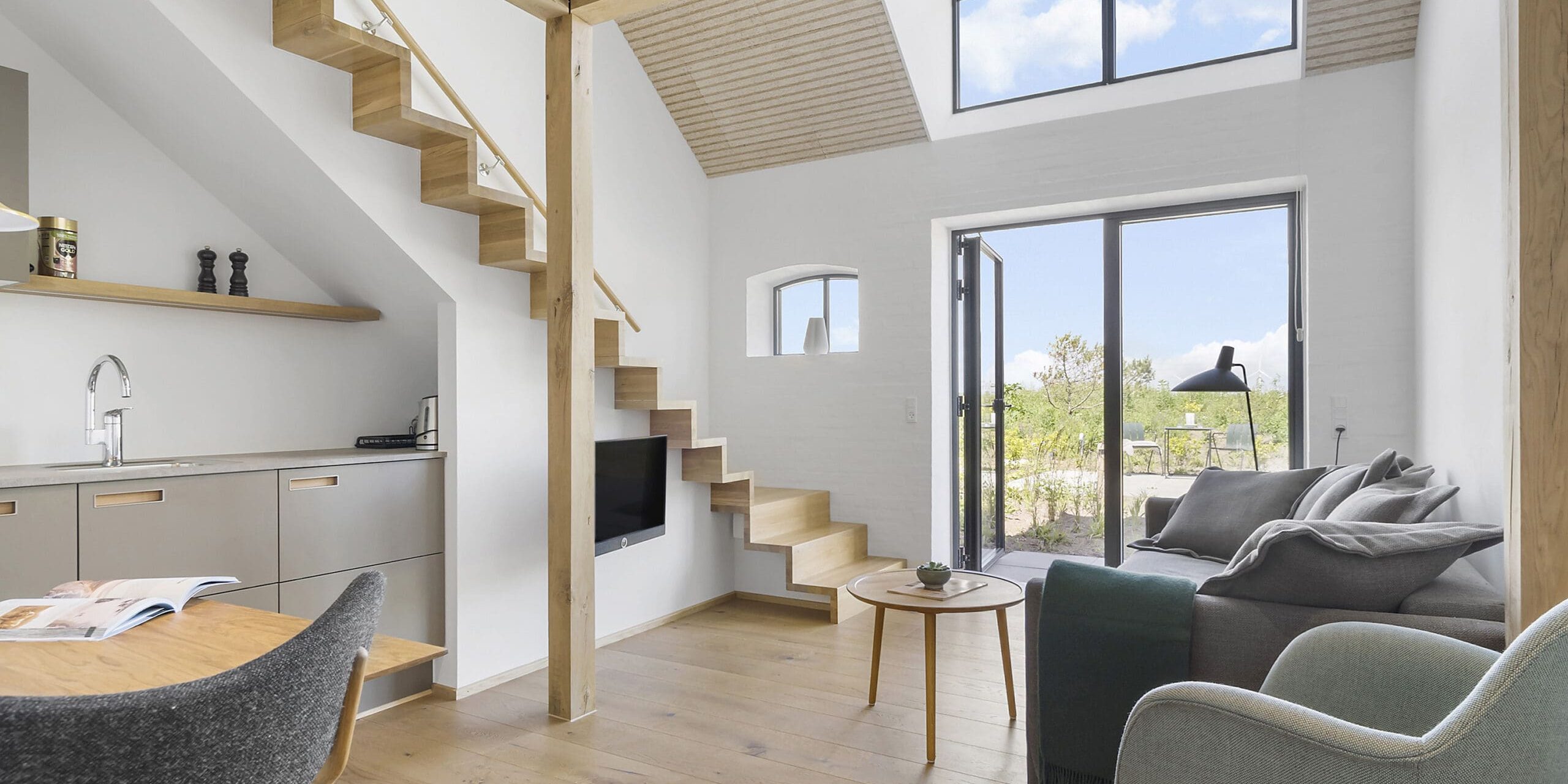 ferielejlighed danmark møllegården ringkøbing fjord Et moderne, minimalistisk opholdsrum med en trætrappe, højt til loftet og store vinduer. Værelset har en grå sofa og lænestol, et lille rundt bord, et fladskærms-tv på en hvid væg og et køkkenområde under trappen. Naturligt lys fylder rummet.