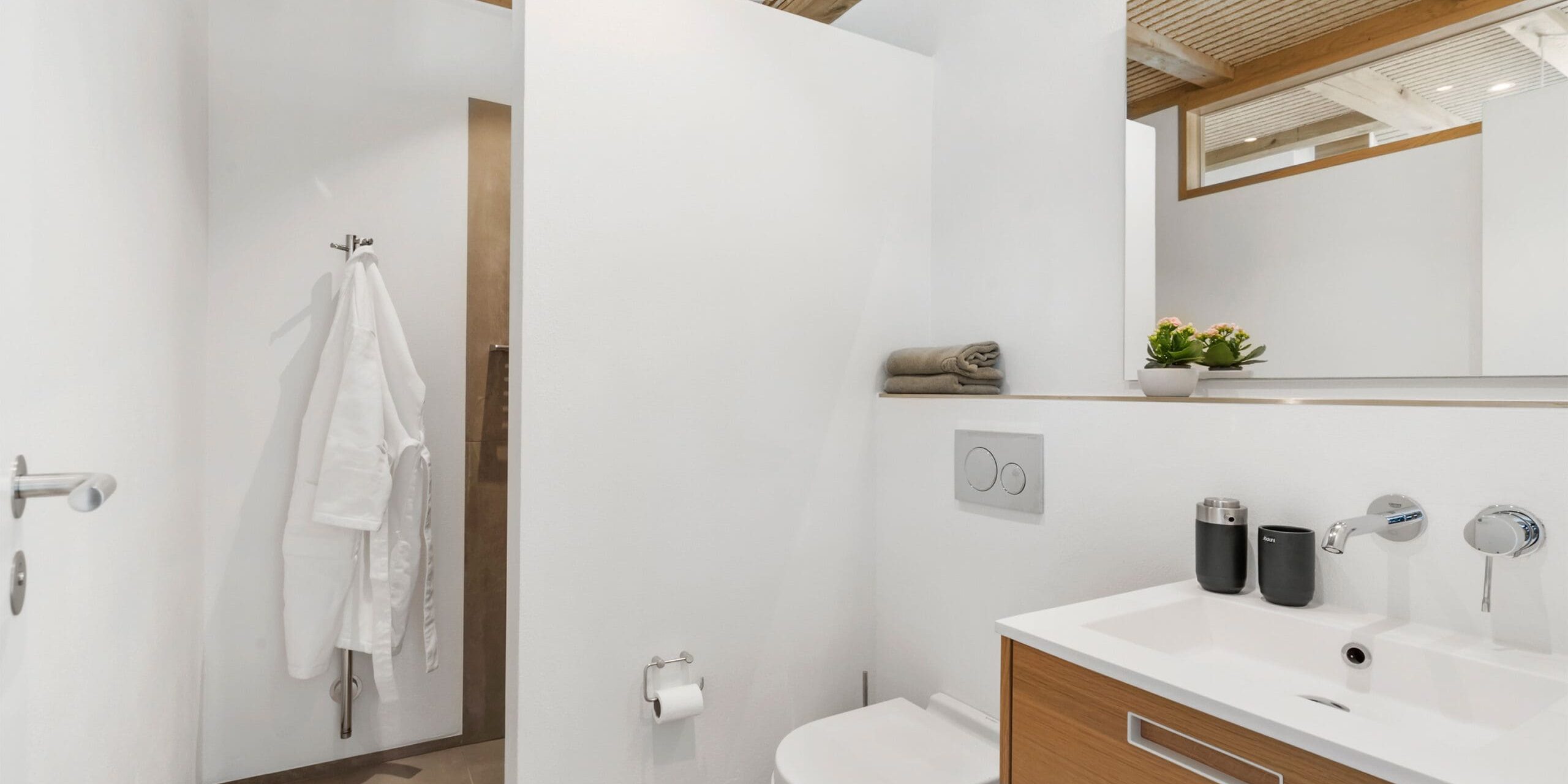 ferielejlighed danmark møllegården ringkøbing fjord Et moderne badeværelse med hvide vægge og træaccenter med et hvidt toilet, en trævask med vask, et stort spejl og en hylde med en potteplante og et foldet håndklæde. Til venstre hænger en badekåbe ved siden af toiletpapirholderen.