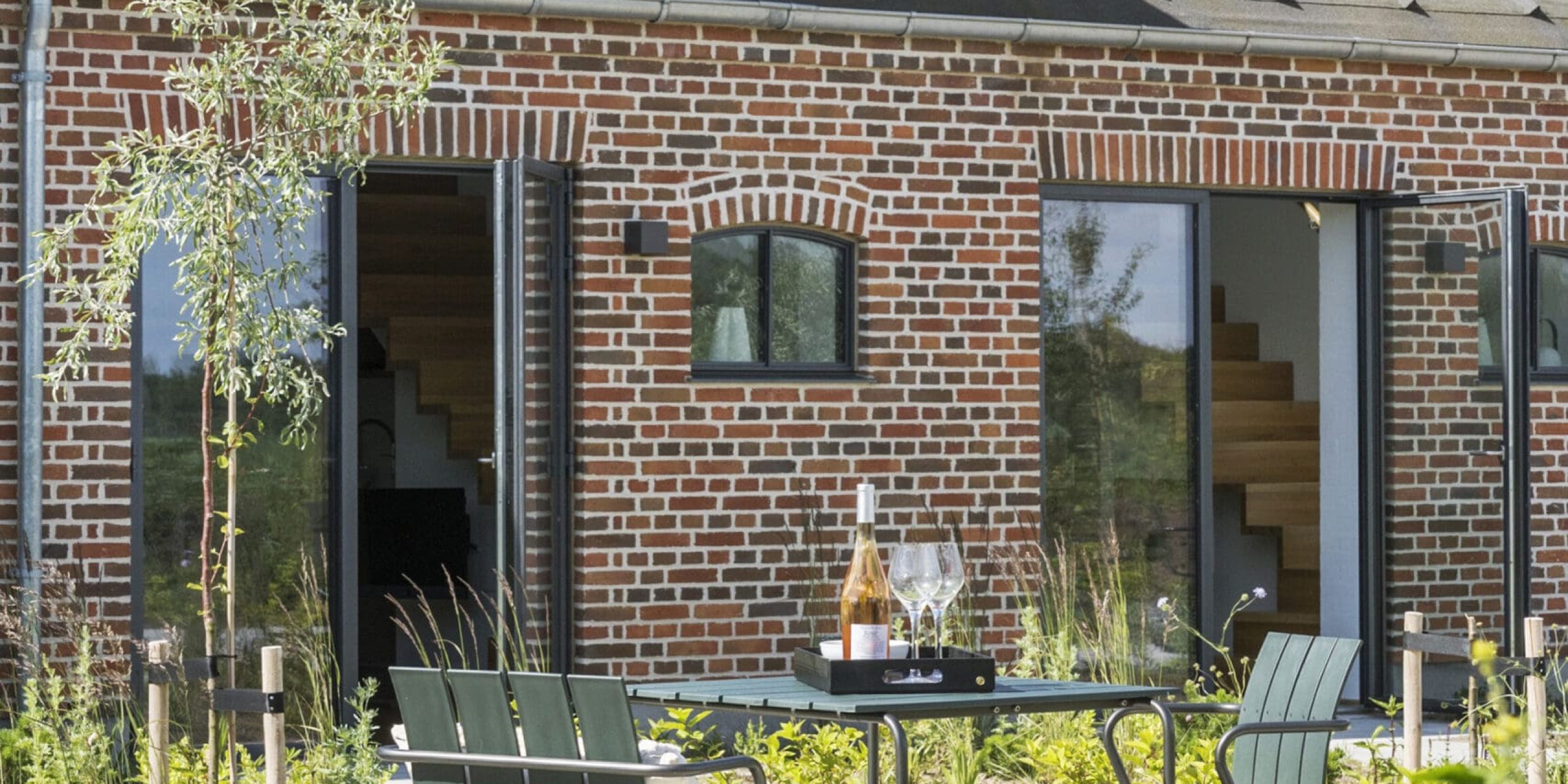 ferielejlighed danmark møllegården ringkøbing fjord Et murstenshus med moderne store vinduer, kviste og skrå tag. I forgrunden er der en udendørs terrasse med grønne stole og et bord med vinflasker og glas. Den omkringliggende have er frodig med forskellige planter og buske. Solrigt vejr.