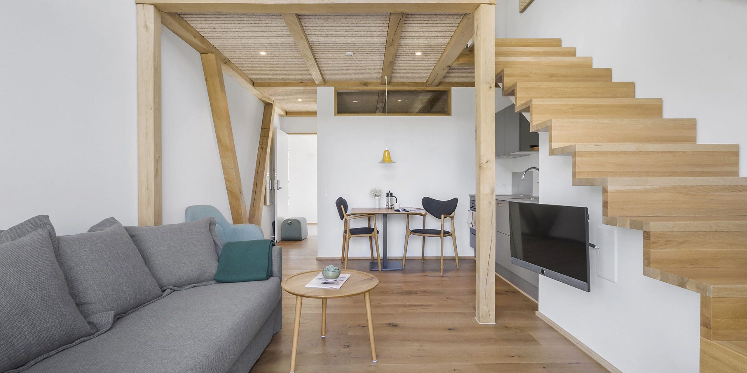 ferielejlighed danmark møllegården ringkøbing fjord Et moderne opholdsrum med trægulv og et åbent design. En grå sofa sidder til venstre, et sofabord af træ foran og en minimalistisk trætrappe uden stiger, der fører til et øverste niveau. Et lille spisebord med stole er bagerst.