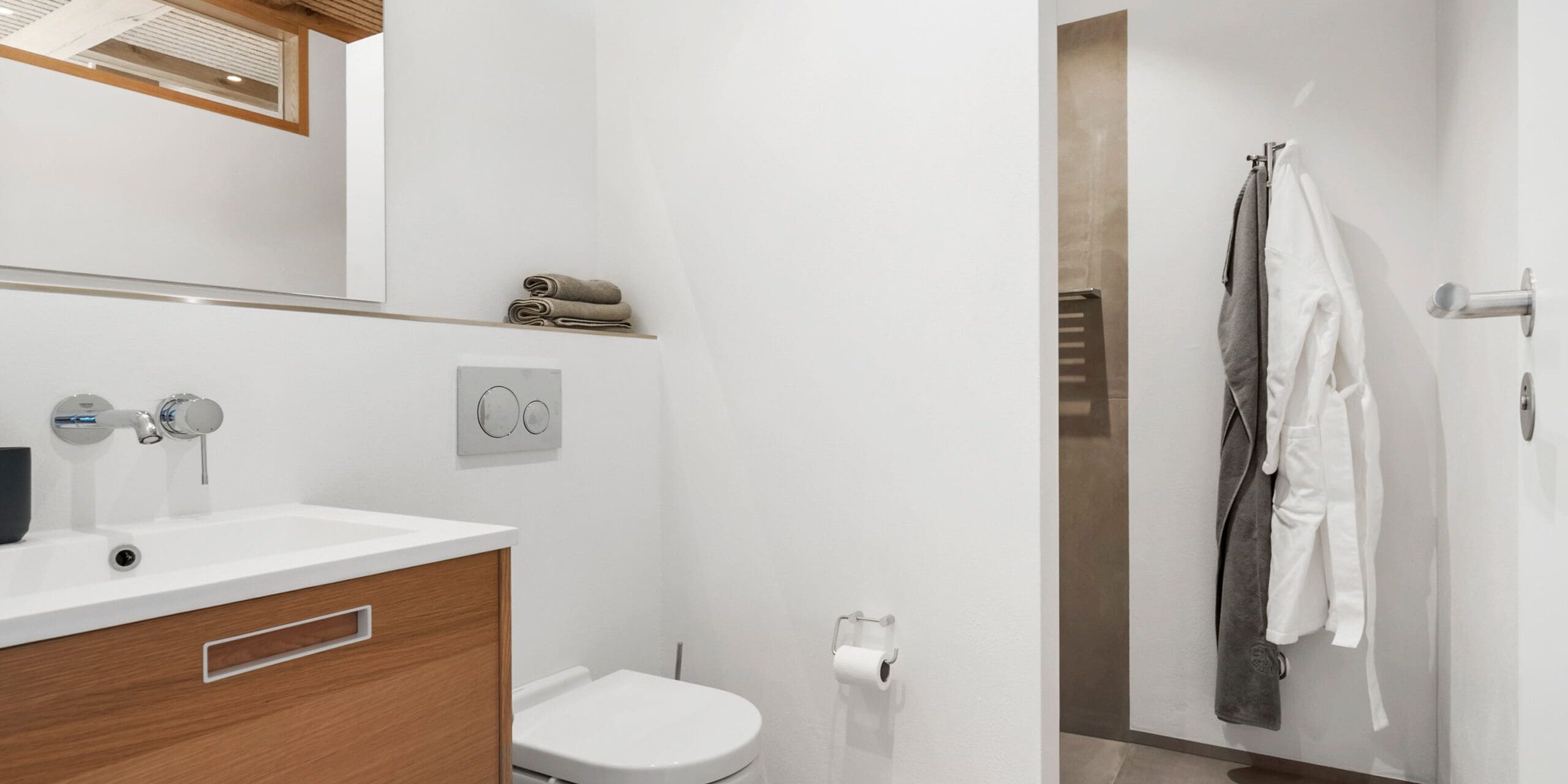 ferielejlighed danmark møllegården ringkøbing fjord Et moderne badeværelse med hvide vægge og træloft. En vask med et træskab, et spejl over og et hvidt toilet er til venstre. Håndklæder er pænt foldet på en hylde over toilettet. Til højre er der et åbent bruseområde med klæder hængende på kroge.