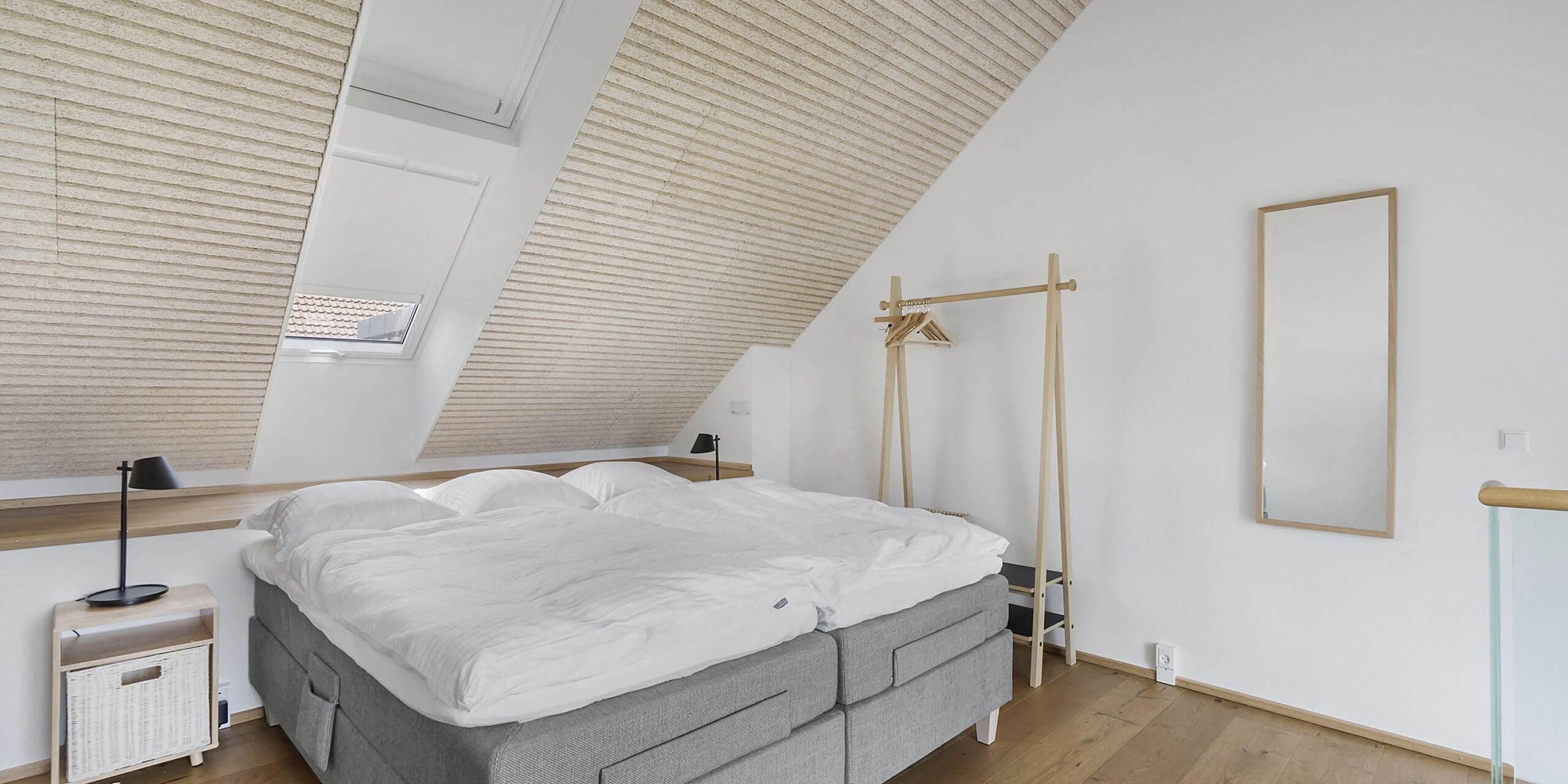 ferielejlighed danmark møllegården ringkøbing fjord Et minimalistisk soveværelse med skråloft har en stor seng med hvidt sengetøj. Værelset inkluderer et selvstændigt tøjstativ i træ, et langt rektangulært spejl, et fletskab og en lille sort lampe. Et ovenlysvindue lukker naturligt lys ind.