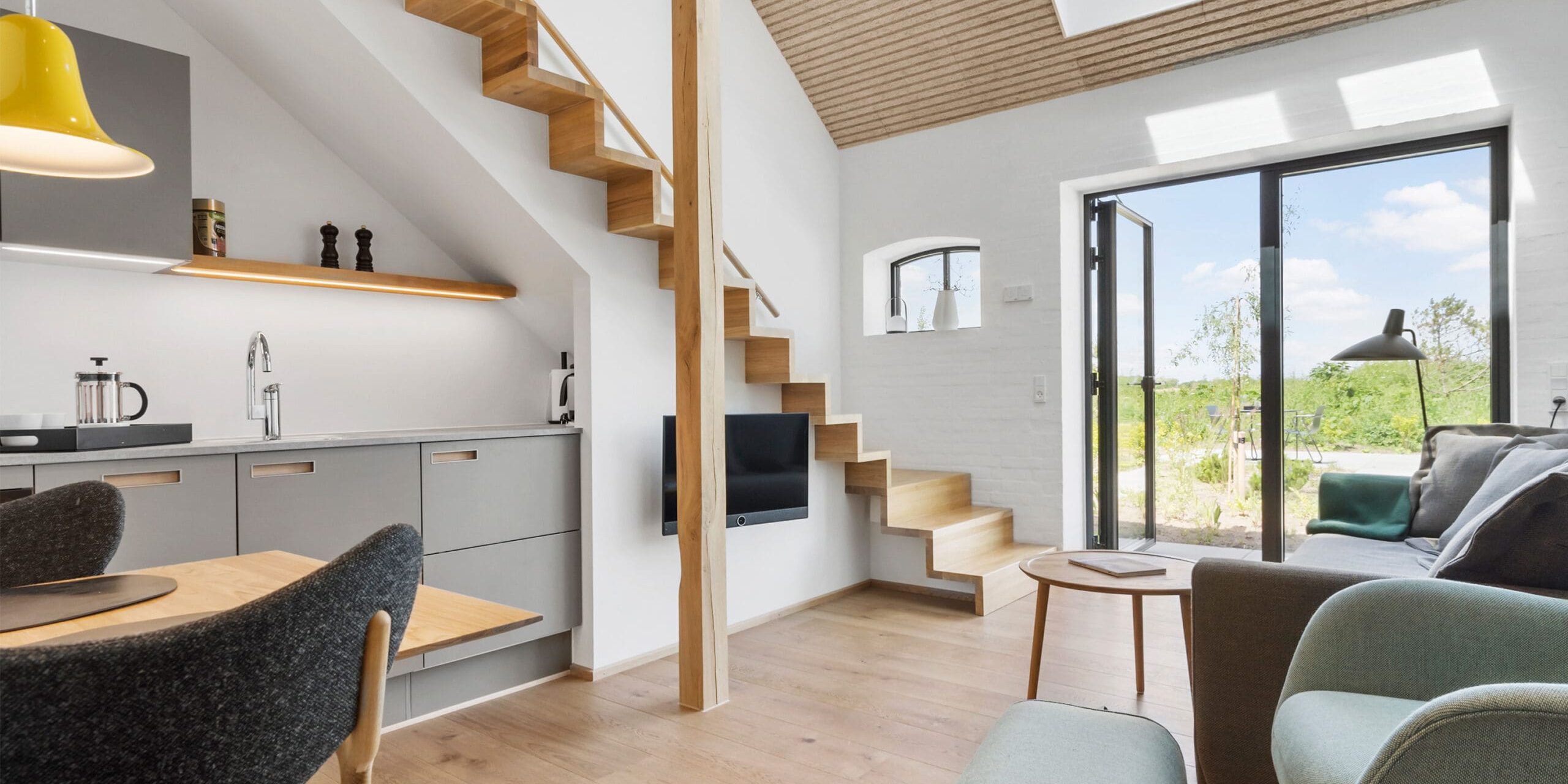 ferielejlighed danmark møllegården ringkøbing fjord Et moderne, lyst opholdsrum med trægulve, en grå sofa og hyggelige stole. Et lille køkkenområde med grå skabe og en gul pendel er til venstre. En trætrappe fører til en hems, og store glasdøre åbner til et solrigt udeområde.