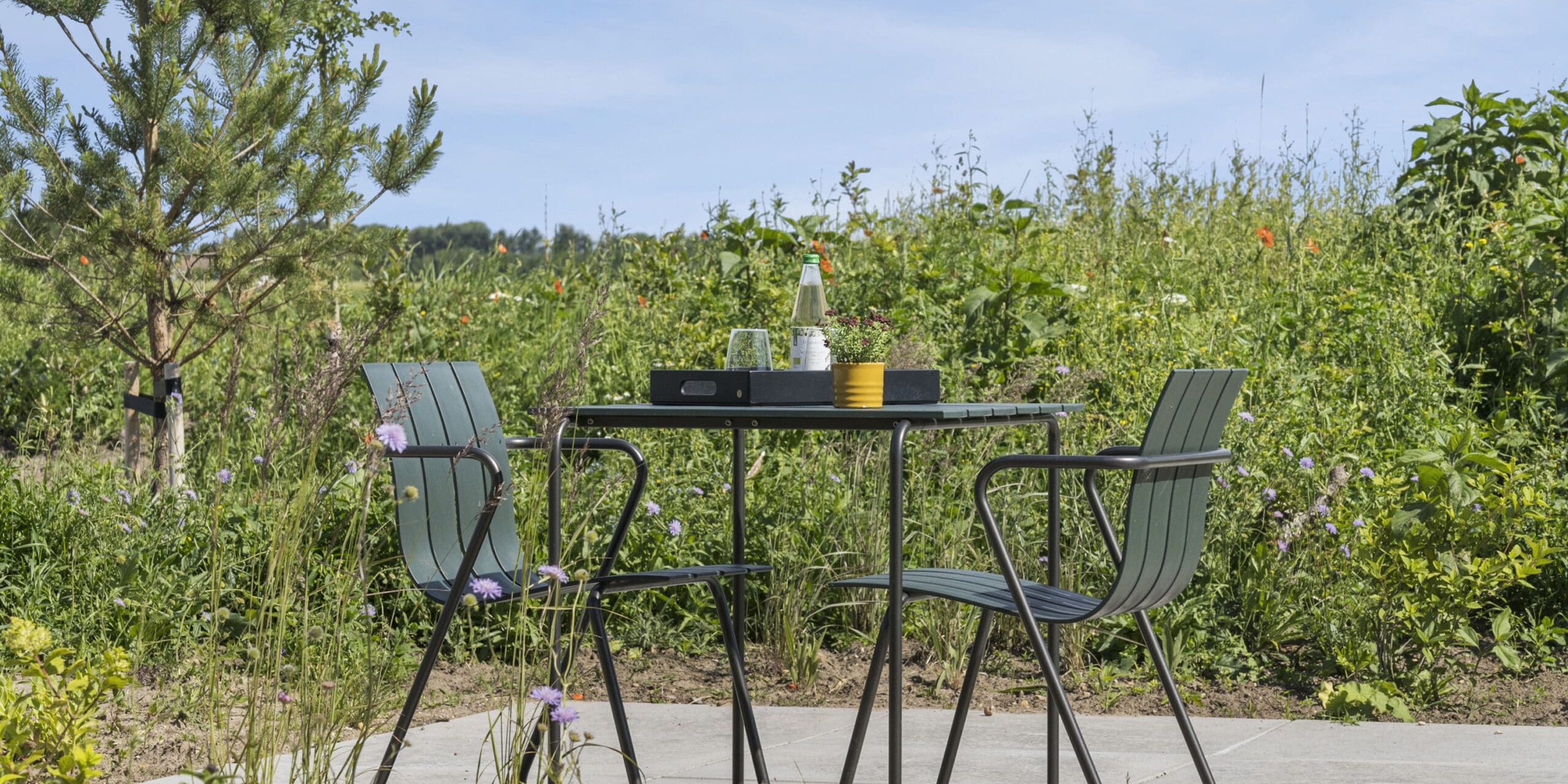 ferielejlighed danmark møllegården ringkøbing fjord En fredfyldt udendørs scene med et lille firkantet bord med to stole. Bordet rummer et gult krus, en flaske og en bakke med småting. Baggrunden er frodig med grønt, blomster og en klar blå himmel, hvilket fremkalder en fredelig og indbydende atmosfære.