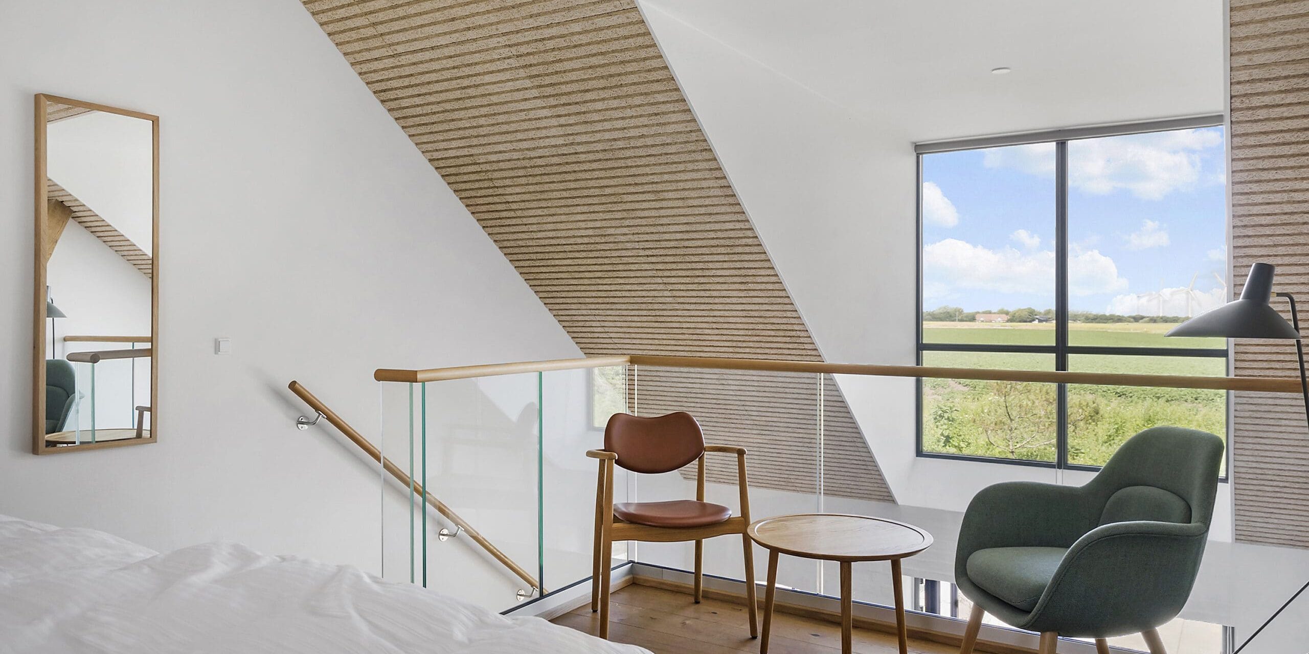 ferielejlighed danmark møllegården ringkøbing fjord Et moderne loftssoveværelse med træloft og et stort vindue med udsigt over landskabet. Værelset er indrettet med en seng, et rundt træbord og to stole - den ene brun og den anden grøn. Et højt spejl hænger på venstre væg, og et glasrækværk bidrager til det åbne design.