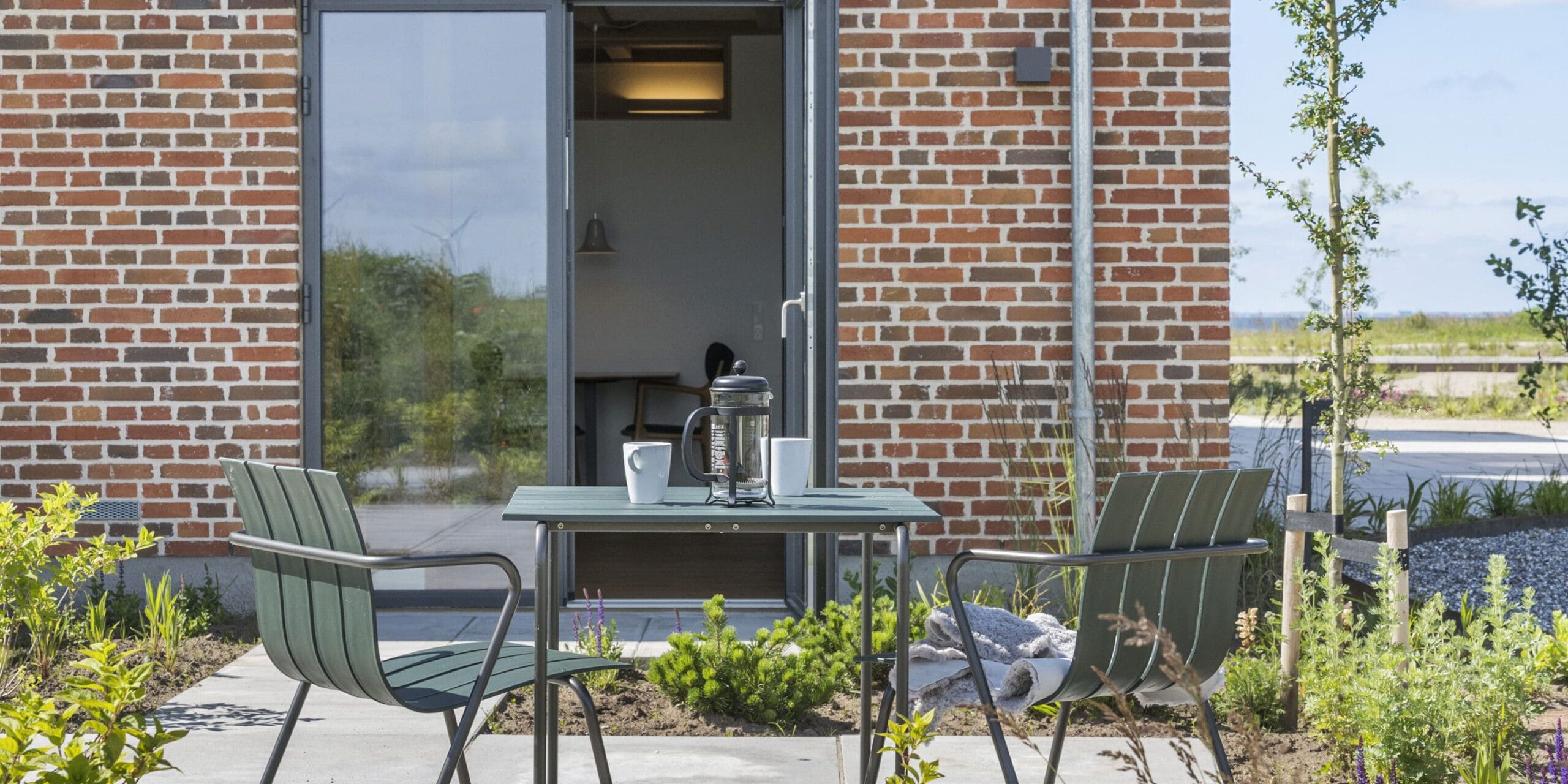 ferielejlighed danmark møllegården ringkøbing fjord En hyggelig gårdhave med et lille bord med en lanterne og to kopper, omgivet af to grønne stole. Terrassen er sat mod en murstensvæg med en glasdør, der fører ind. Scenen er udsmykket med planter og har udsigt over et naturskønt, åbent landskab.