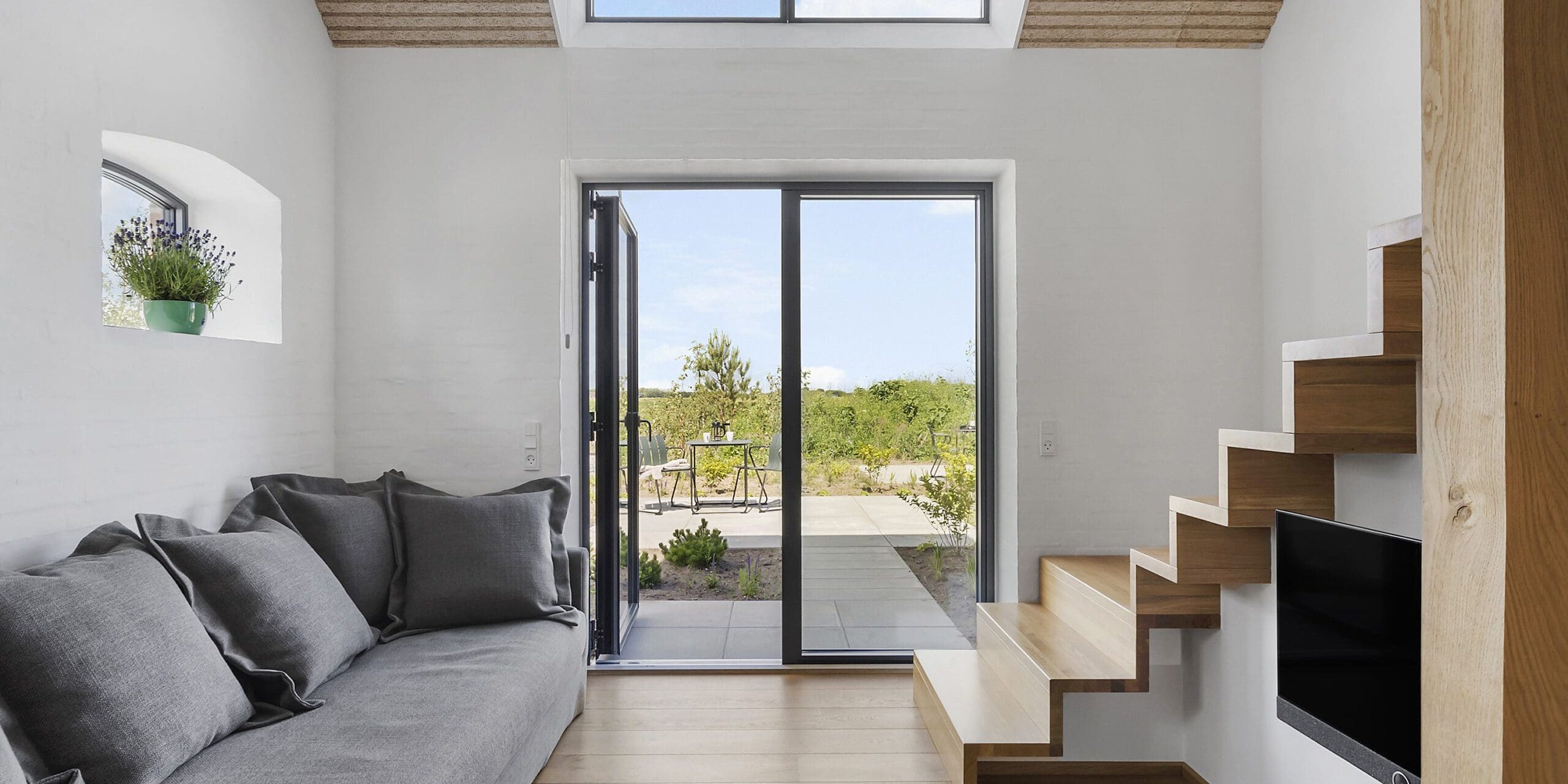 ferielejlighed danmark møllegården ringkøbing fjord En moderne, minimalistisk stue med hvide vægge og trægulve. Den har en grå sofa, potteplanter, et tv og en unik flydende trappe. Store glasskydedøre åbner ud til en terrasse med havemøbler og udsigt til grønne områder under en klar blå himmel.