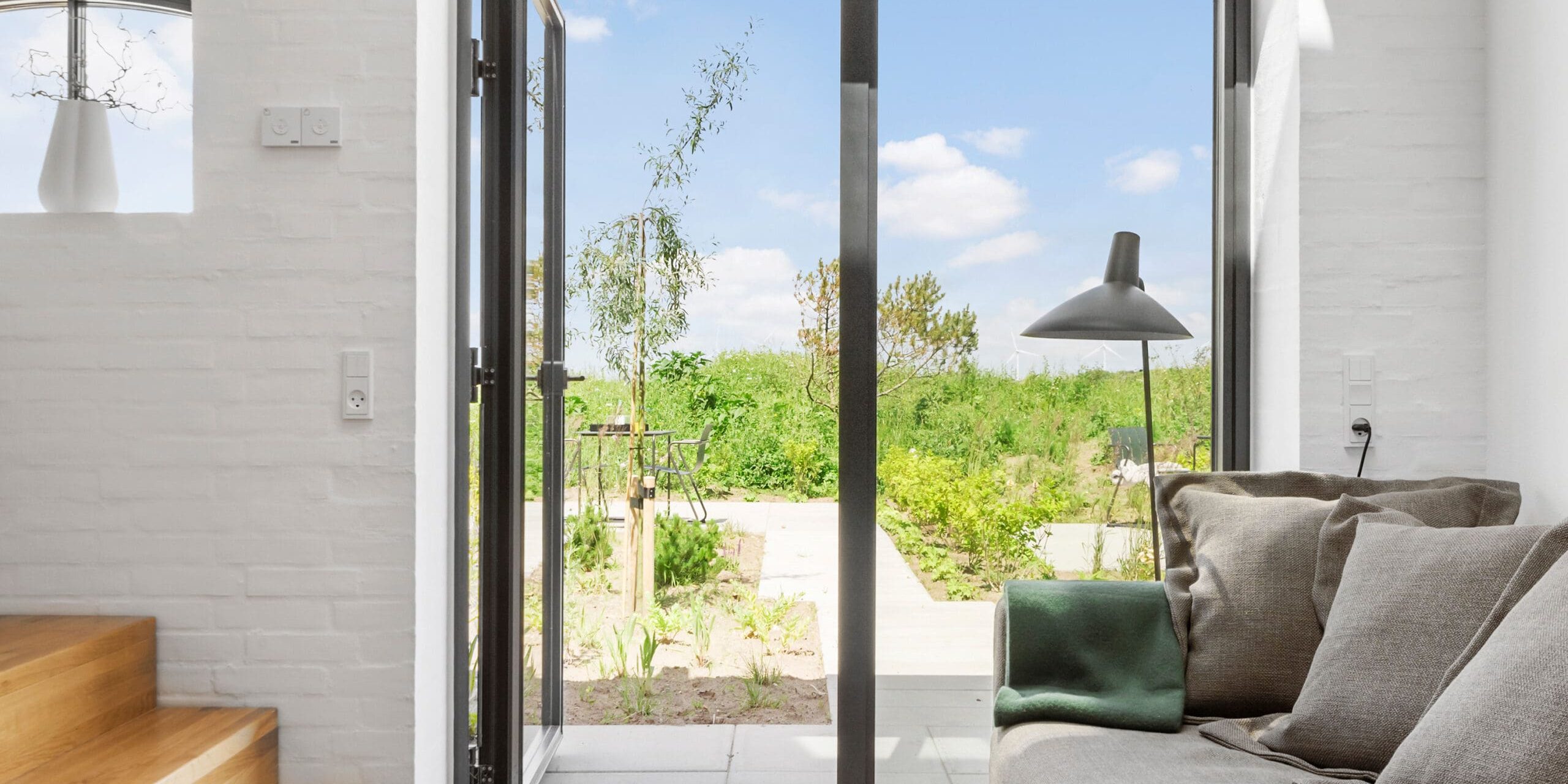 ferielejlighed danmark møllegården ringkøbing fjord Et lyst, moderne opholdsrum med en glasdør, der fører til en frodig have. Interiøret har en grå sofa med hynder, en elegant gulvlampe og trætrapper ved siden af en hvid murstensvæg. Naturligt lys oversvømmer rummet og skaber en luftig og indbydende atmosfære.