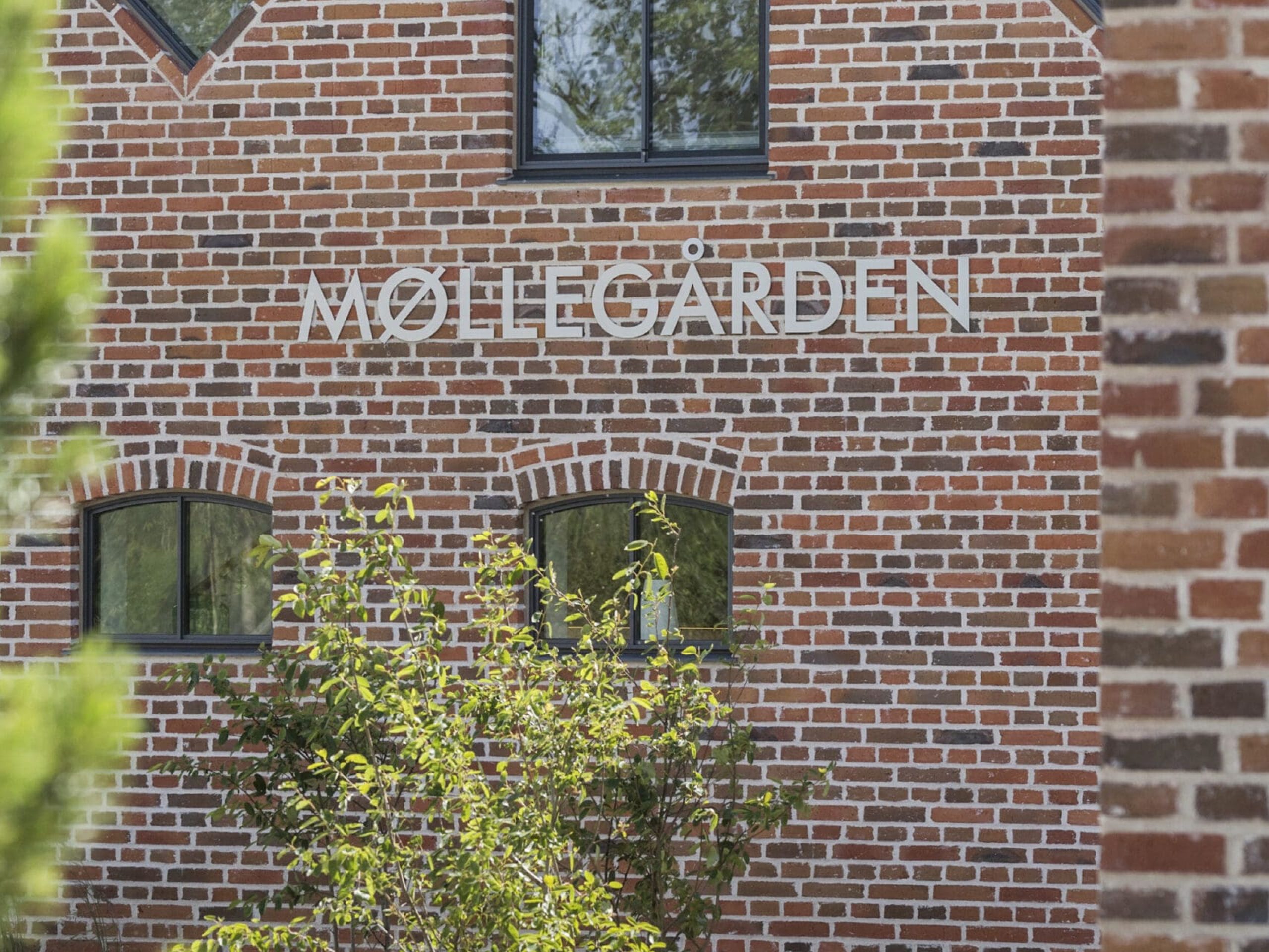 ferielejlighed danmark møllegården ringkøbing fjord En murstensbygning med navnet "MØLLEGÅRDEN" vist med hvide bogstaver. Bygningen har mørkrammede vinduer og er delvist skjult af en mindre murstenskonstruktion i forgrunden. Der er grønt, herunder buske og græs, foran bygningen.