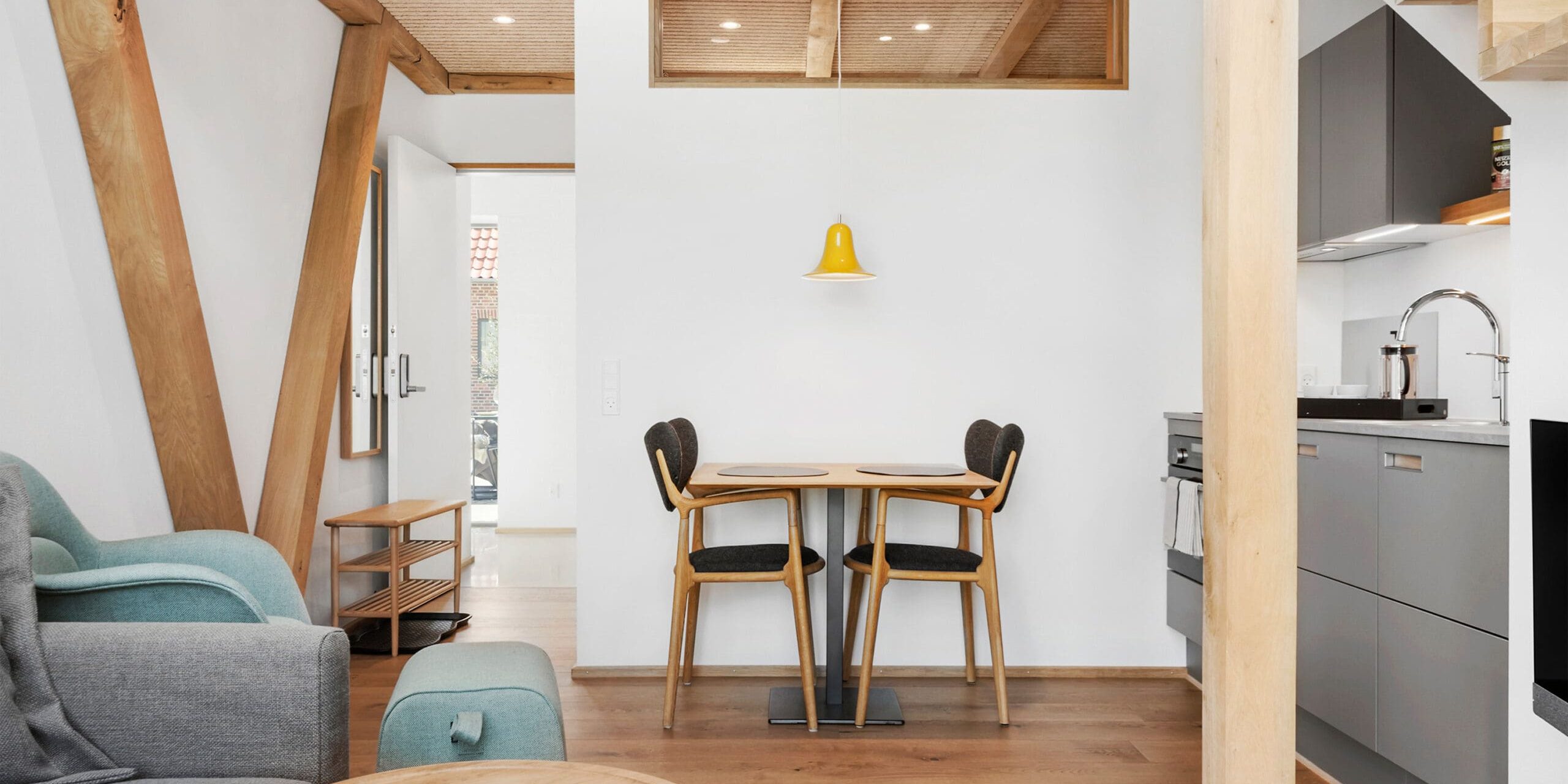ferielejlighed danmark møllegården ringkøbing fjord En moderne, minimalistisk opholds- og spisestue med træbjælker og gulv. Rummet omfatter et lille rundt spisebord med to sorte stole, en grå sofa, et sofabord i træ og et køkken med grå skabe. En gul pendel hænger over spisebordet.