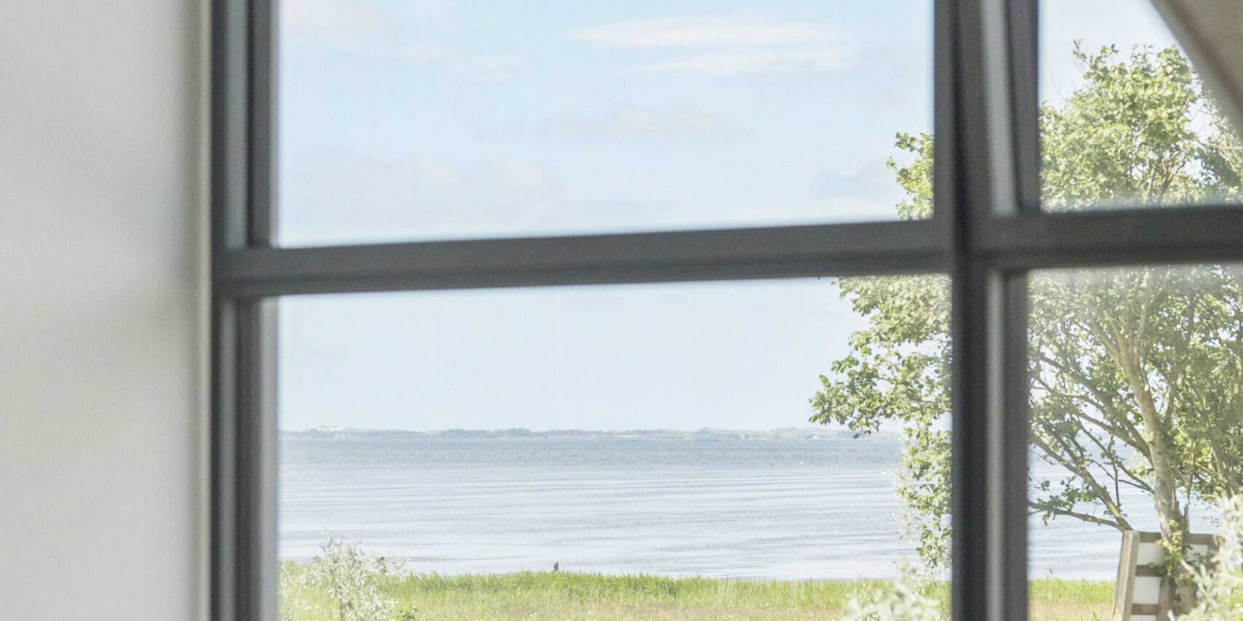 ferielejlighed danmark møllegården ringkøbing fjord En fredfyldt udsigt over en rolig sø er synlig gennem et stort gulv-til-loft-vindue. Forgrunden viser et minimalistisk interiør med rullede yogamåtter på gulvet, og udenfor har en bænk udsigt over det fredelige vand og grønne områder. Himlen er delvist overskyet.