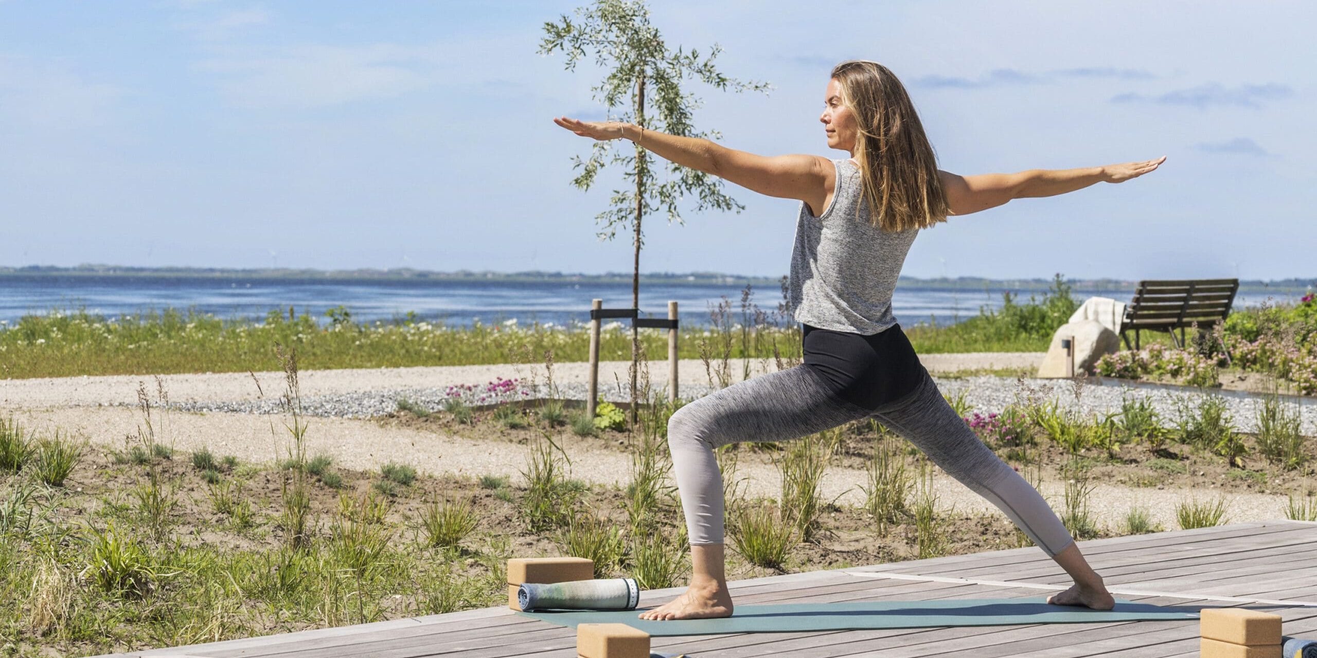 ferielejlighed danmark møllegården ringkøbing fjord En kvinde dyrker yoga i en krigerstilling på et trædæk med udsigt over et fredfyldt kystlandskab. Yogamåtter og -klodser er placeret ved siden af hende. Scenen er rolig og solrig med udsigt over havet, sandede stier, grønne områder og en bænk i baggrunden.