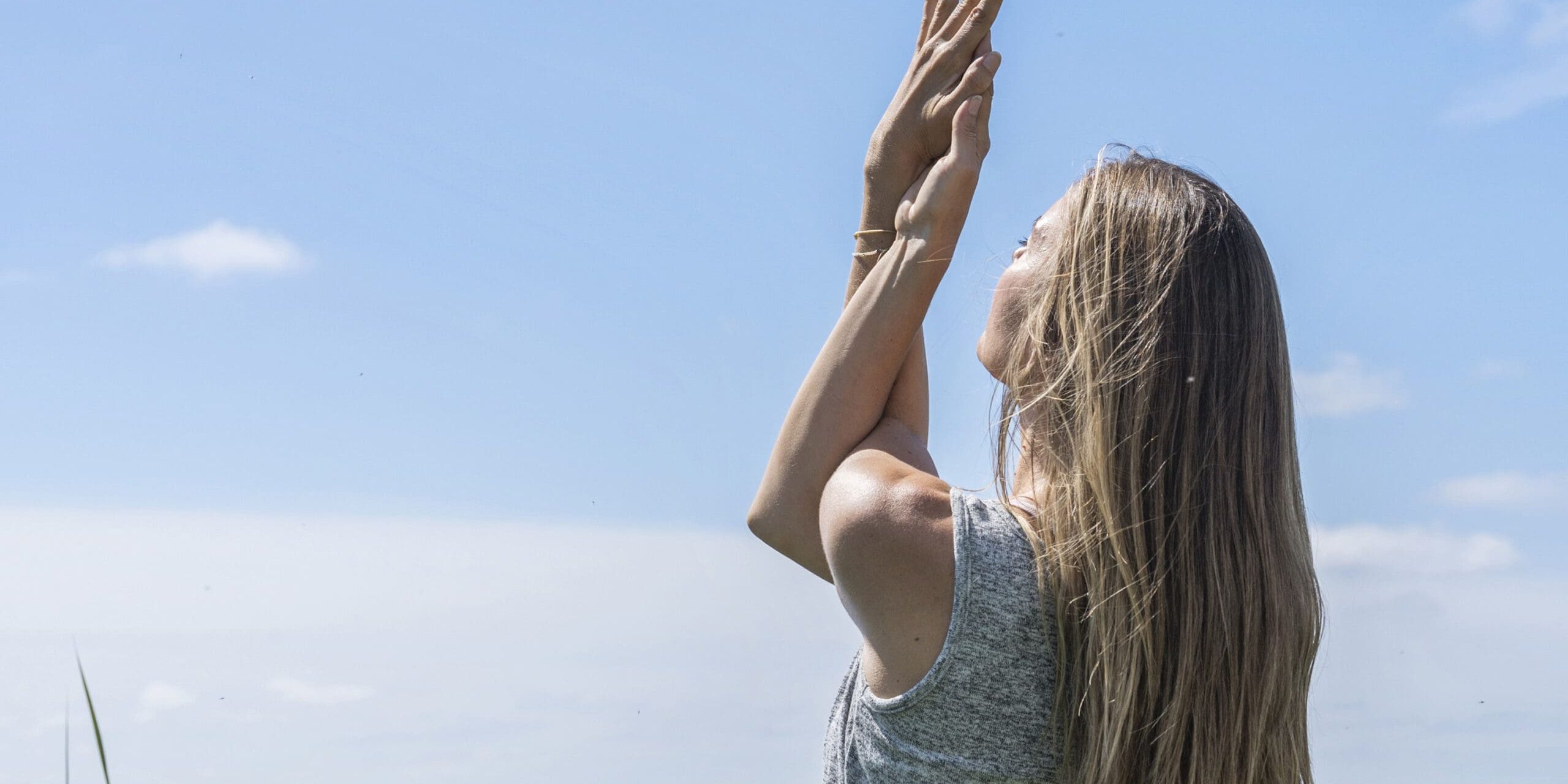 ferielejlighed danmark møllegården ringkøbing fjord En kvinde med langt blondt hår står nær en vandmasse og rækker armene op mod himlen i en yogastilling. Hun er iført en lysegrå ærmeløs top, og himlen er klar og blå med enkelte spredte skyer.