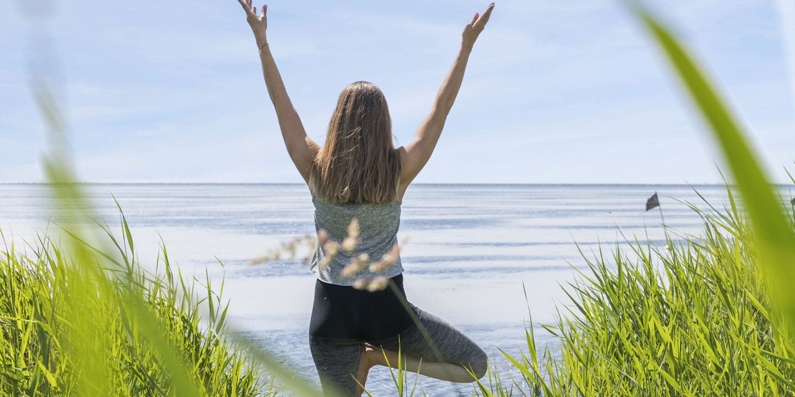 ferielejlighed danmark møllegården ringkøbing fjord En person, der står nær højt græs ved vandet, vender væk med armene løftet. De balancerer på ét ben i en yogastilling mod en fredfyldt baggrund af havet og den blå himmel.