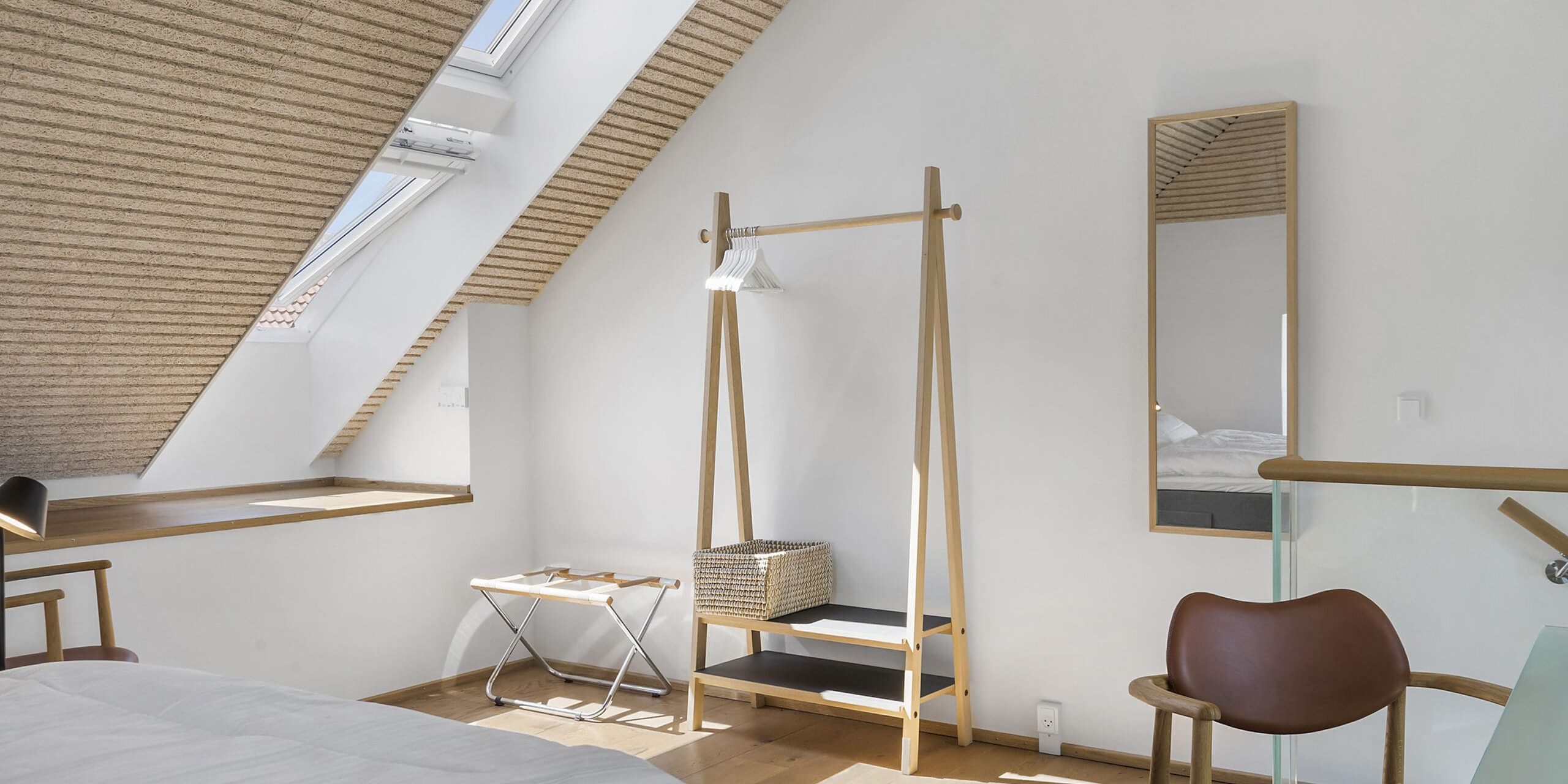 ferielejlighed danmark møllegården ringkøbing fjord Et lyst, minimalistisk loftsværelse med loft til kip og ovenlys. Værelset har en træseng, en lille bænk med en kuffert, et tøjstativ i stigestil med en vævet kurv, et stort spejl og en brun stol. Vægge og sengetøj er hvide.