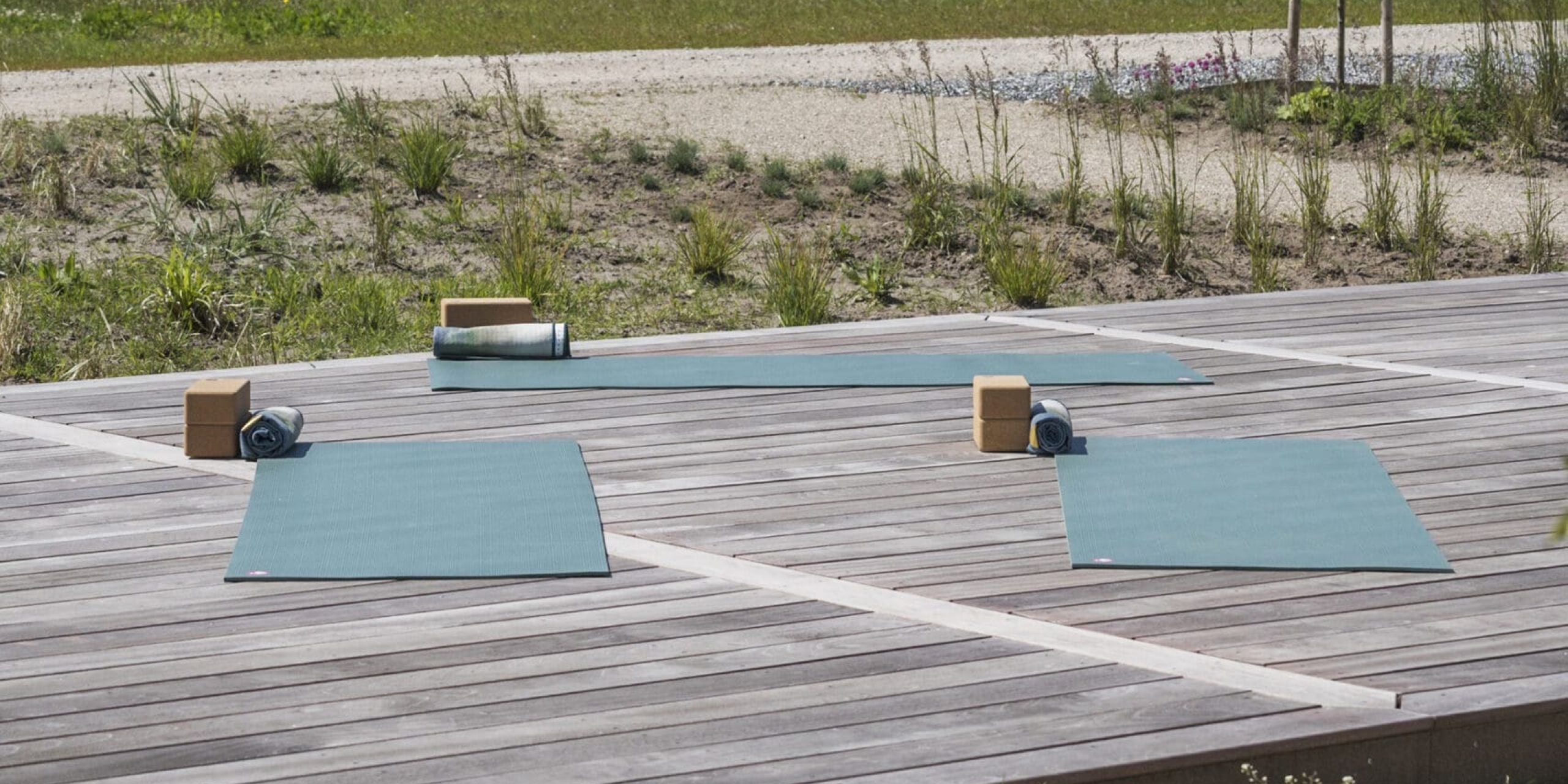 ferielejlighed danmark møllegården ringkøbing fjord To yogamåtter er lagt ud på et trædæk nær kysten sammen med blokke og sammenrullede håndklæder. Ud over dækket er der en sandet sti, der fører til græsklædte marker og et roligt, stort vandområde under en blå himmel med spredte skyer.