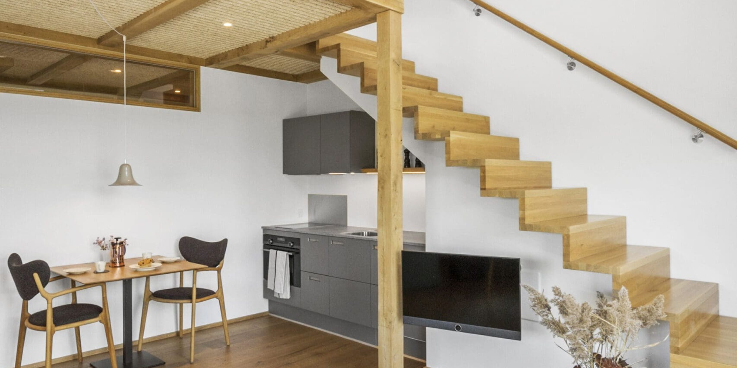 ferielejlighed danmark møllegården ringkøbing fjord Et moderne, minimalistisk opholdsrum i åbent plan med trægulve, en flydende trætrappe med glasrækværk, et lille spisebord med to stole og et kompakt køkken med grå skabe. Ovenpå har en hems med træbjælker udsigt over området.