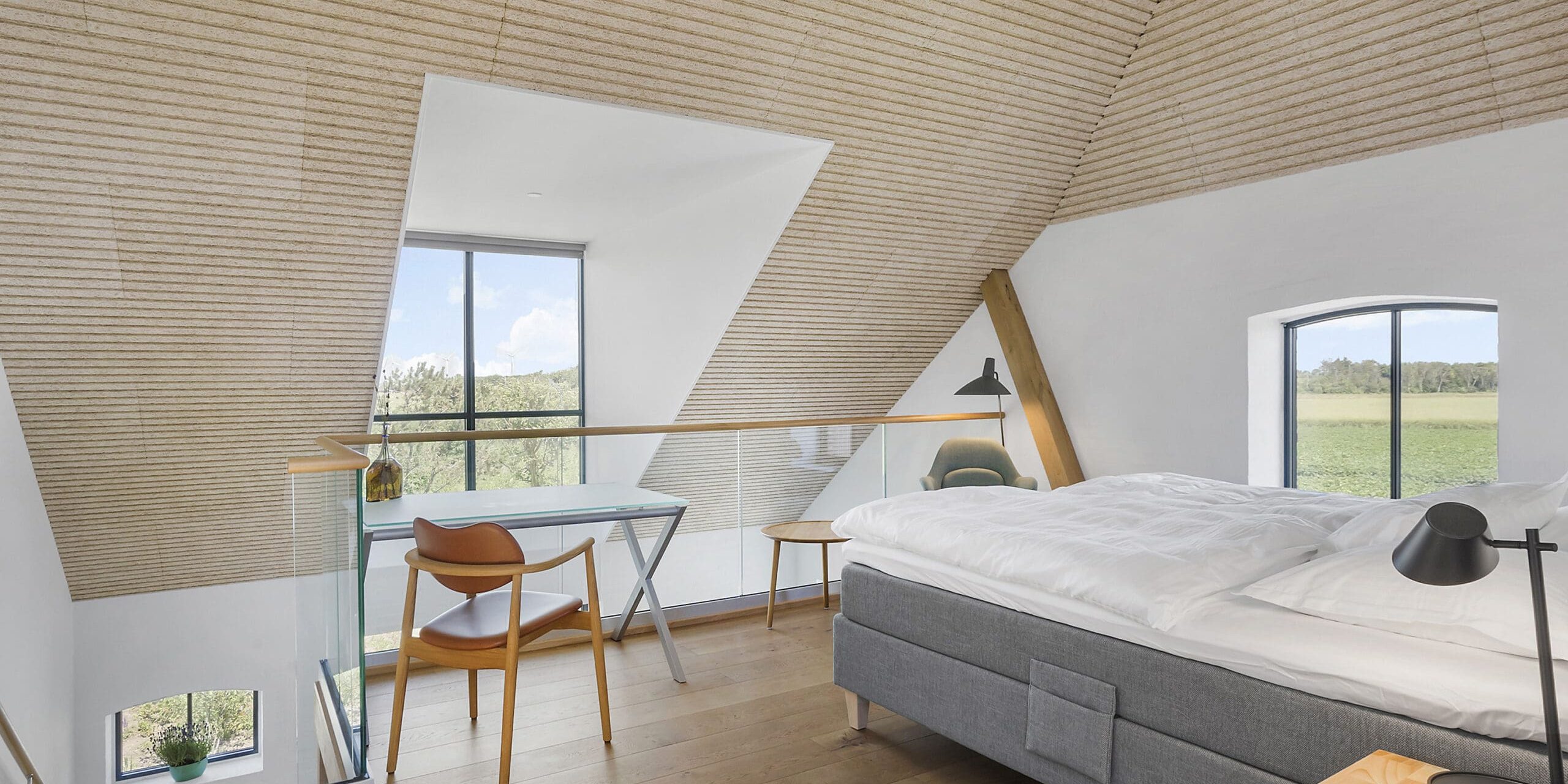 ferielejlighed danmark møllegården ringkøbing fjord Et hyggeligt loftssoveværelse med skrånende træloft og store trekantede vinduer med udsigt til grønne omgivelser udenfor. Værelset har en grå polstret seng, et lille træskrivebord med en bærbar computer, en træstol og en moderne gulvlampe. Naturligt lys fylder rummet.