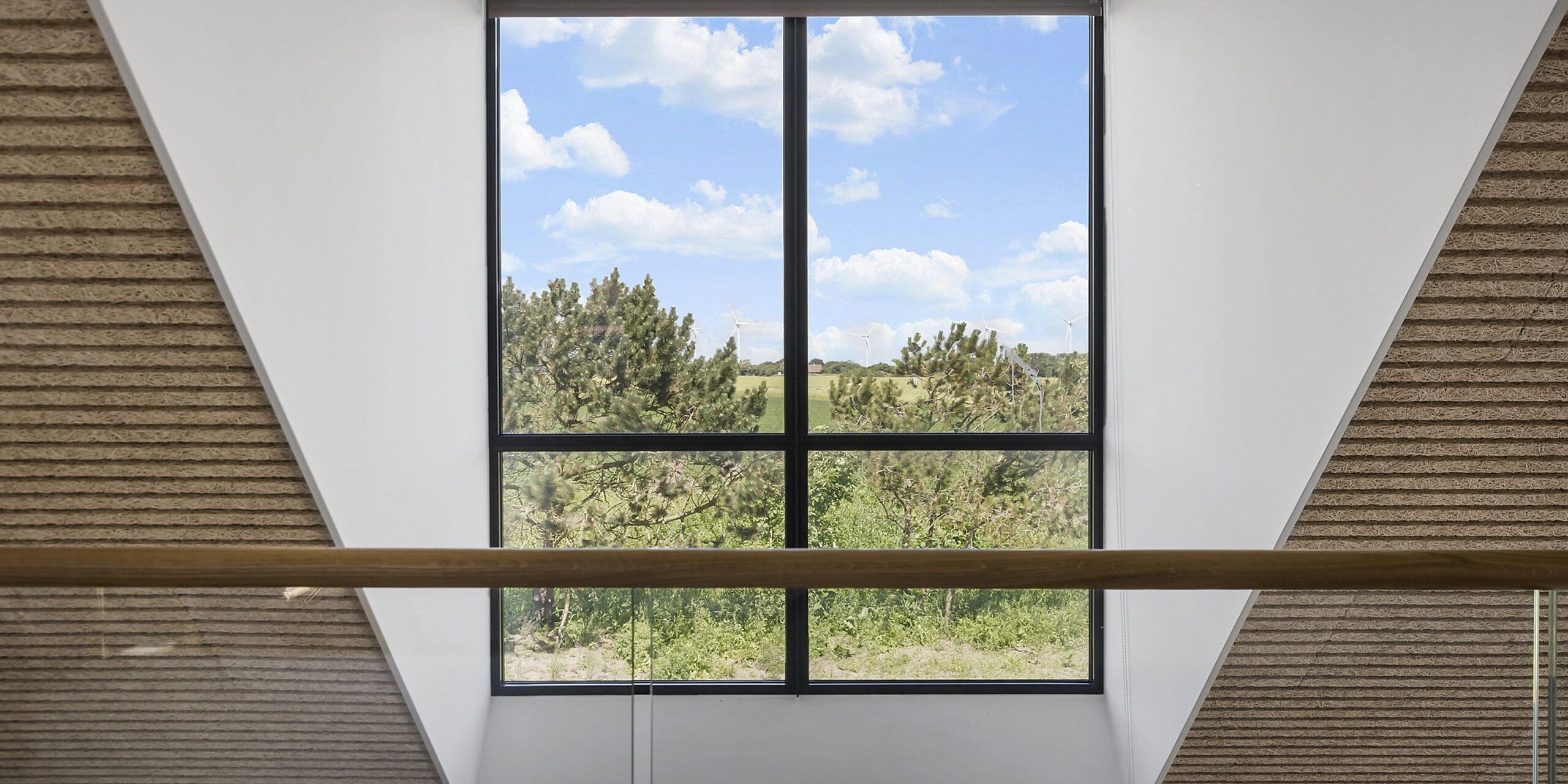ferielejlighed danmark møllegården ringkøbing fjord Et stort, rektangulært vindue med et gittermønster giver en naturskøn udsigt over grønne områder og træer under en blå himmel med bløde hvide skyer. Interiøret byder på moderne, minimalistisk design med rene linjer og neutrale toner, herunder et trægelænder i forgrunden.