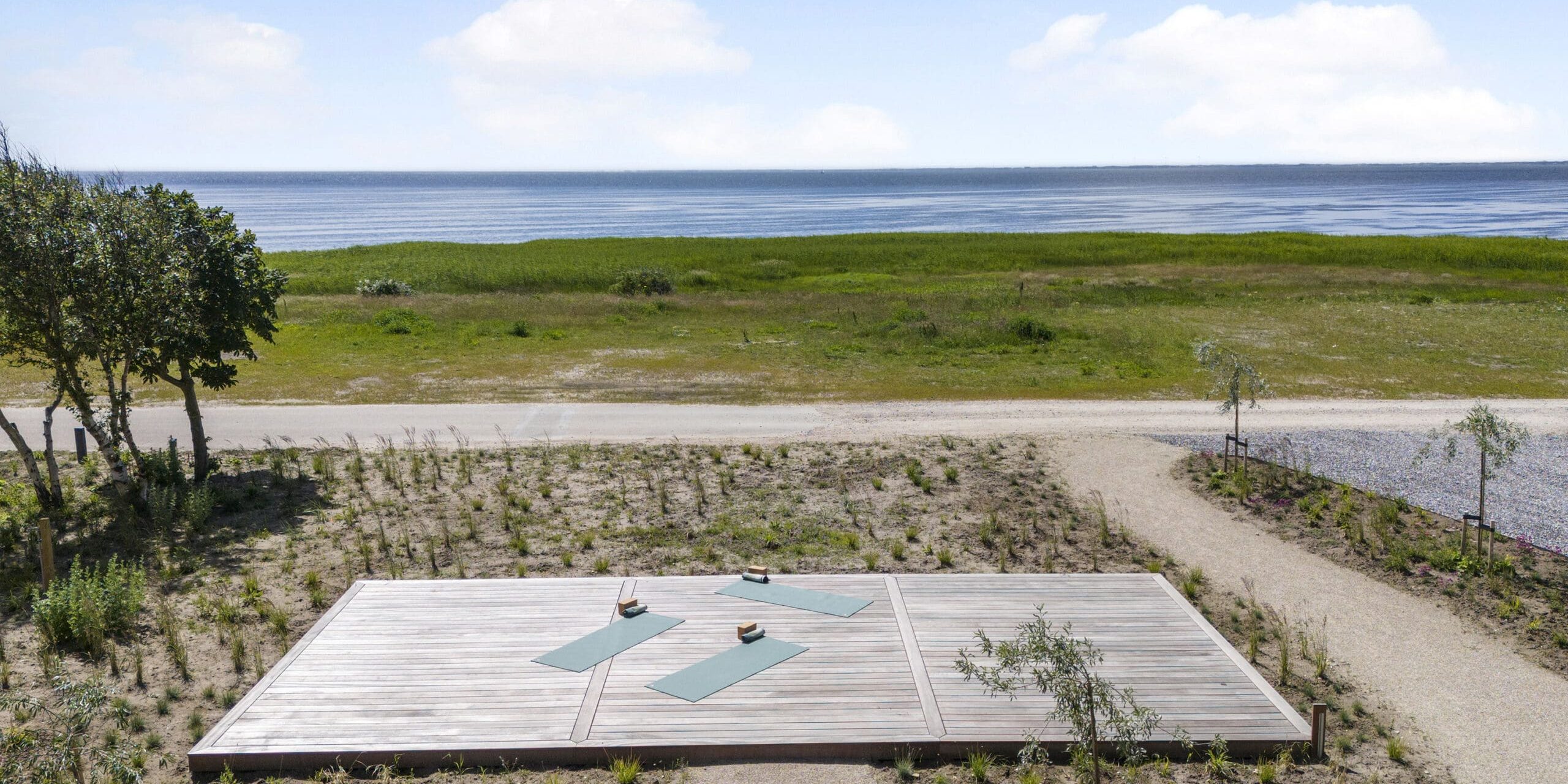 ferielejlighed danmark møllegården ringkøbing fjord En træplatform med tre yogamåtter og to vandflasker er beliggende i naturlige omgivelser omkranset af sand, græs og træer. Ud over platformen fører en sti til et åbent græsareal og en rolig vandmasse, der strækker sig ud til horisonten under en blå himmel.