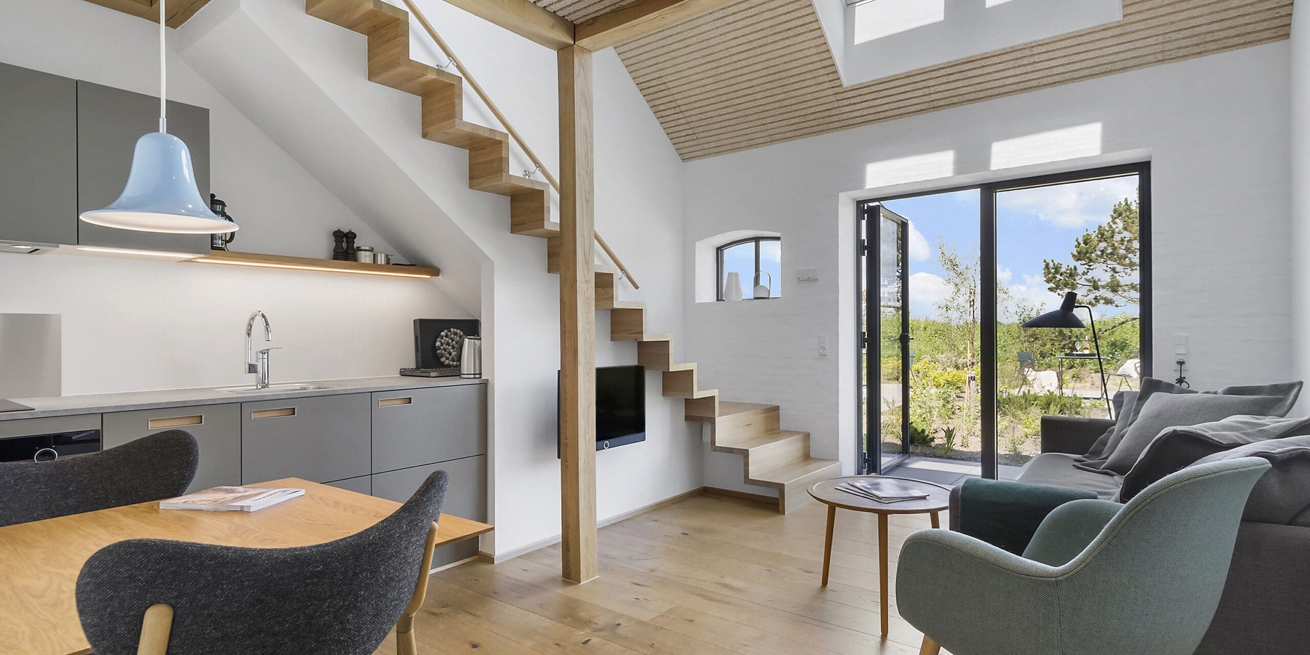 ferielejlighed danmark møllegården ringkøbing fjord Et moderne opholdsrum med åbent koncept med hems. Funktionerne omfatter trætrapper, ovenlys, store vinduer, en grå sofa, grønne og sorte loungestole, et spisebord i træ med sorte stole og et minimalistisk køkken med grå skabe. Lyse trægulve overalt.
